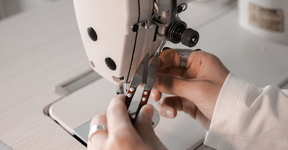 Manos enhebrando tela en una máquina de coser blanca sobre una mesa de costura.