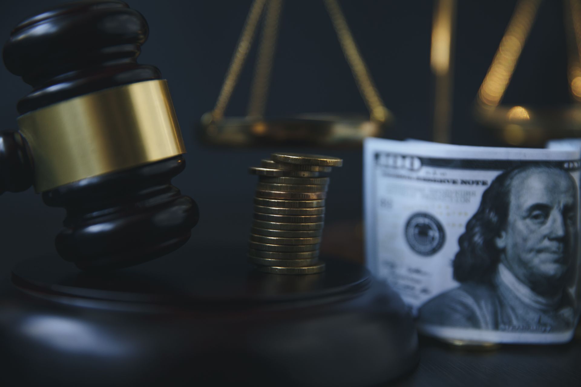 Gavel, scales of justice, stack of coins, and a hundred-dollar bill on a dark surface.