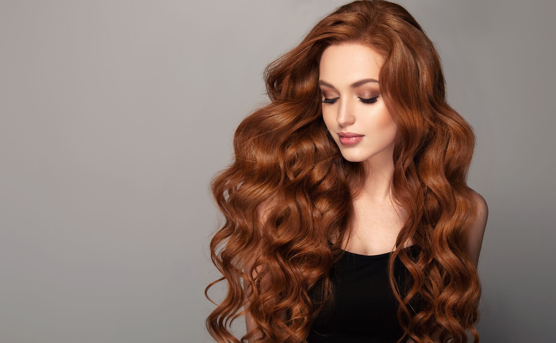Woman with flowing, wavy auburn hair. Wearing a black top against a gray background, looking down.