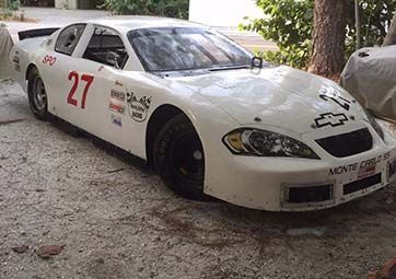 A white race car is parked in a driveway next to a tree.