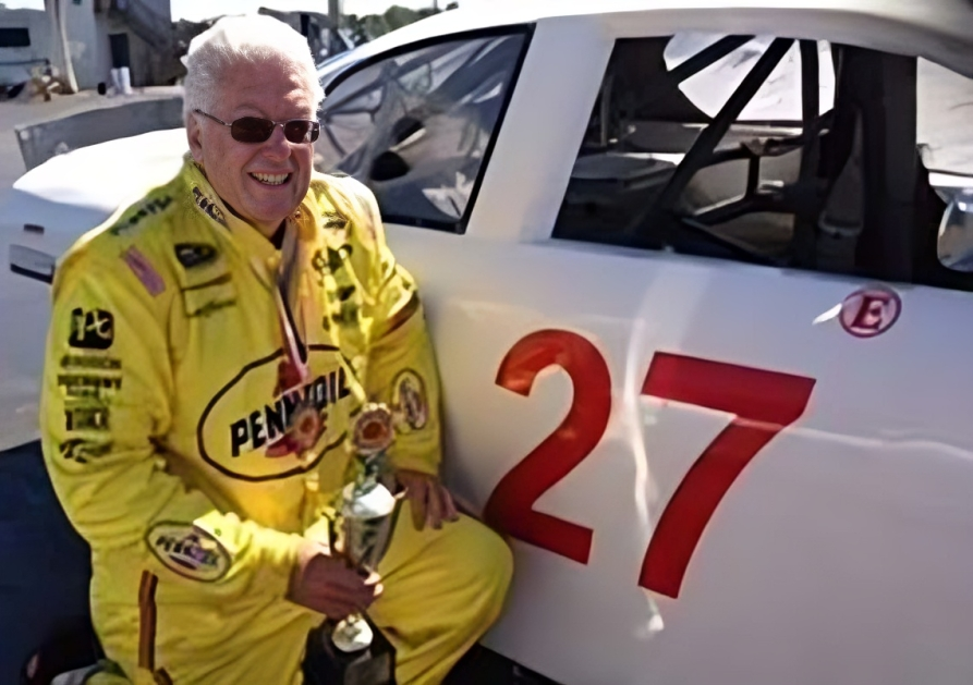 A man in a pennzoil uniform holds a trophy in front of a race car