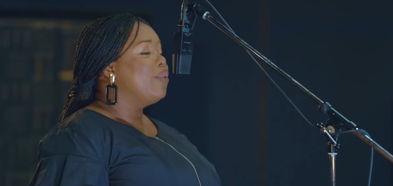 Chevelle singing into a microphone in a dark studio. She wears a black top and earrings.