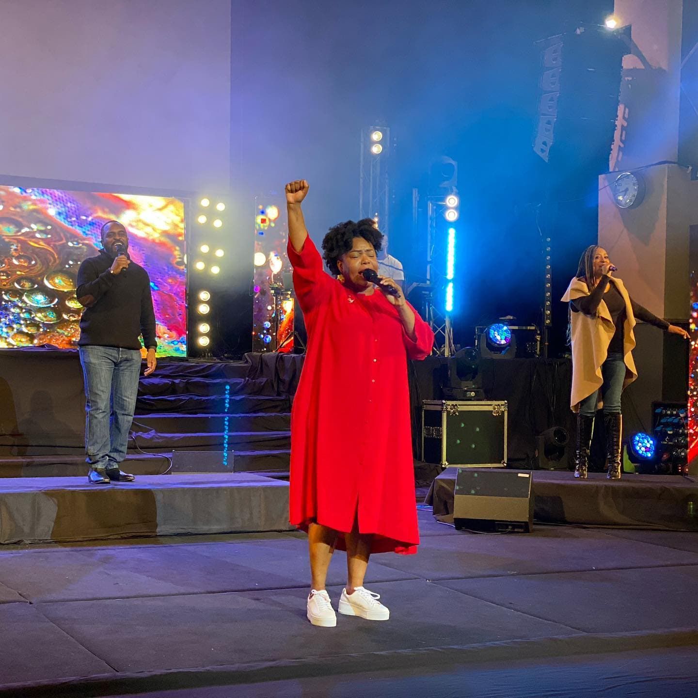  Chevelle in red dress with fist raised, singing onstage with two others.