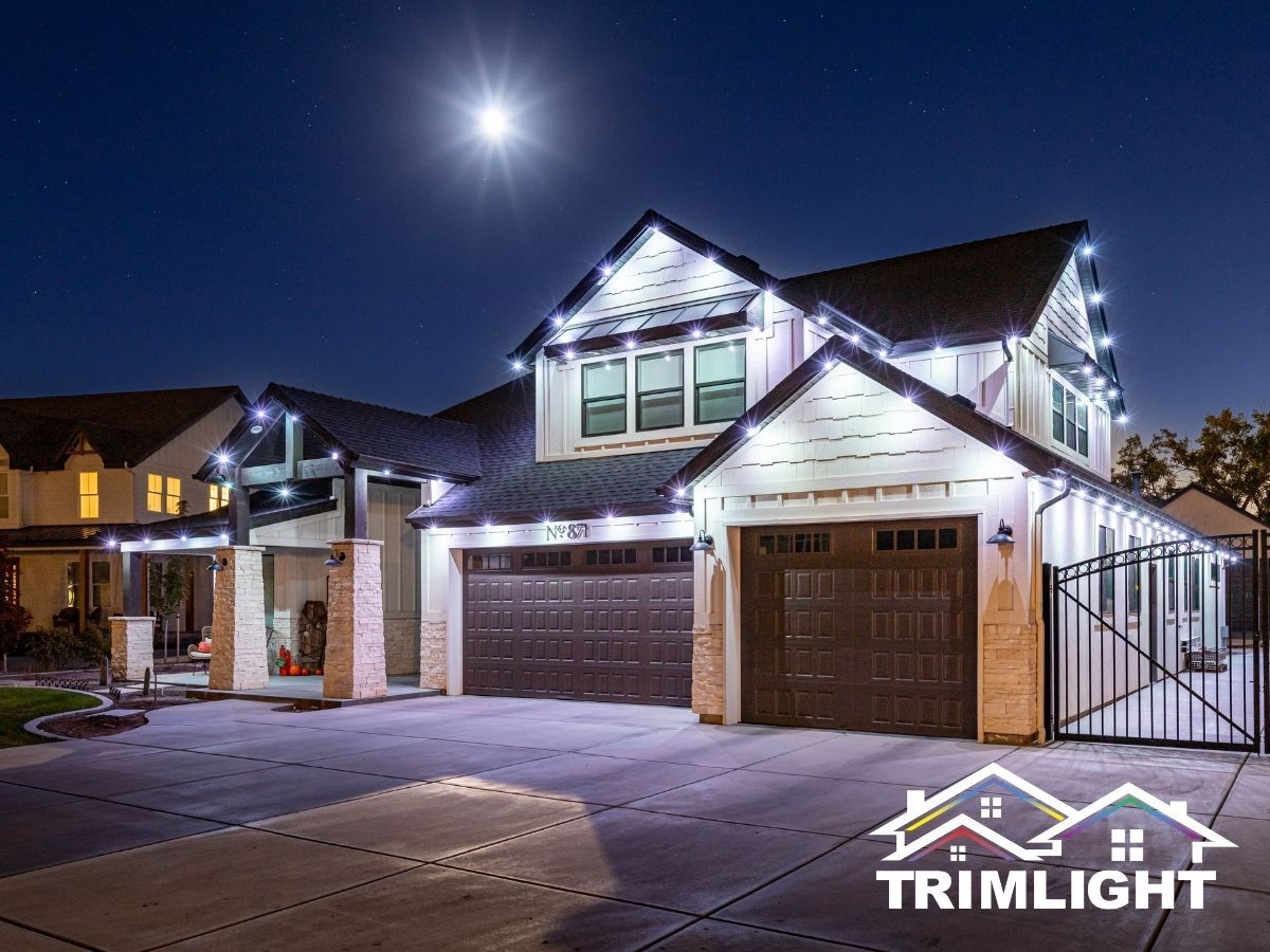 House at night with white trim lights. Brown garage doors, bright moon, and logo in corner.