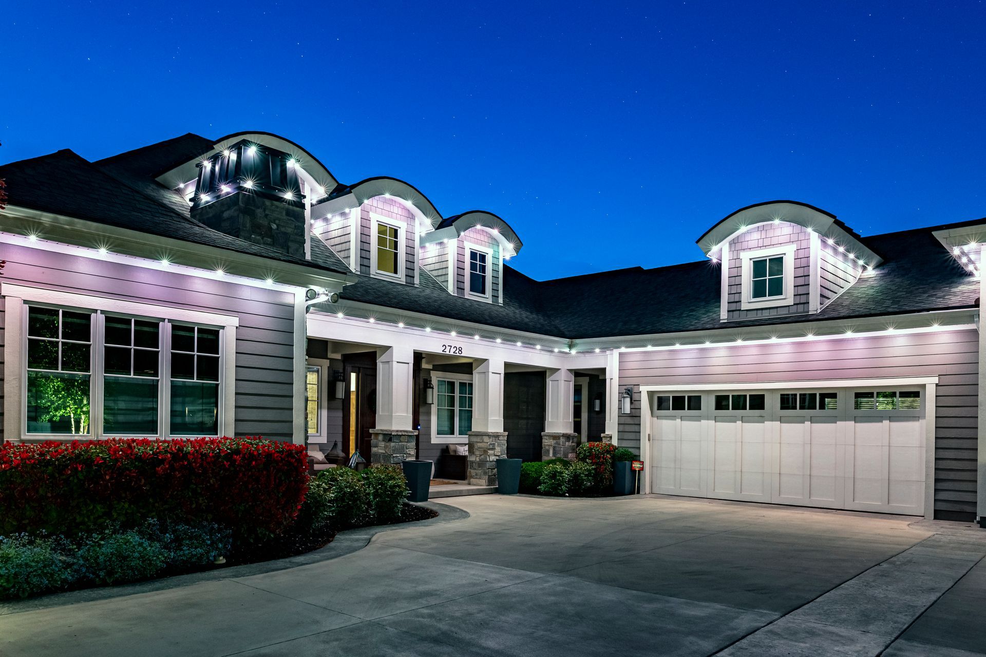A house at night with white lights outlining the roof and windows; driveway and garage.