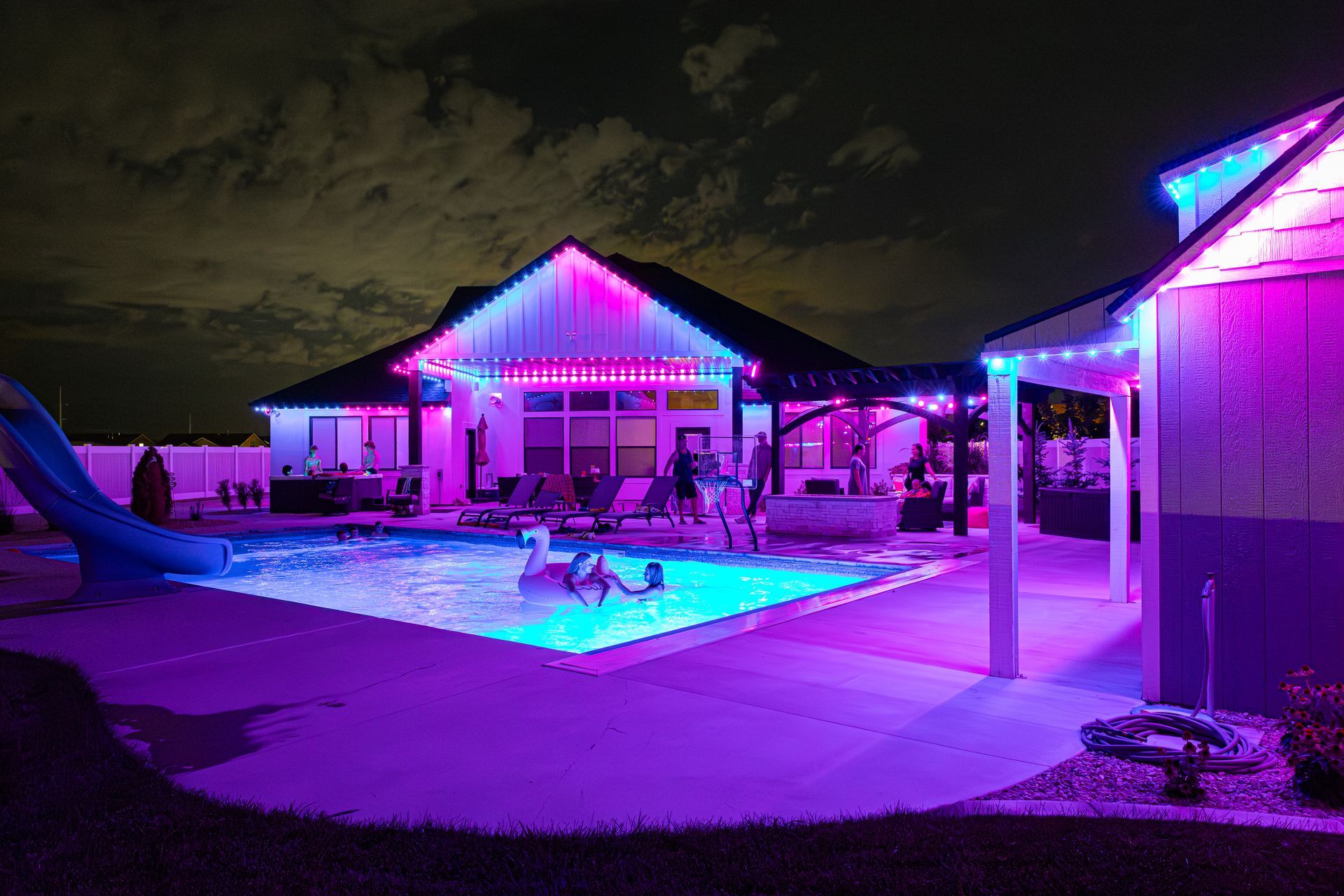Nighttime backyard scene with a pool, house, and pergola illuminated by pink and blue lights.