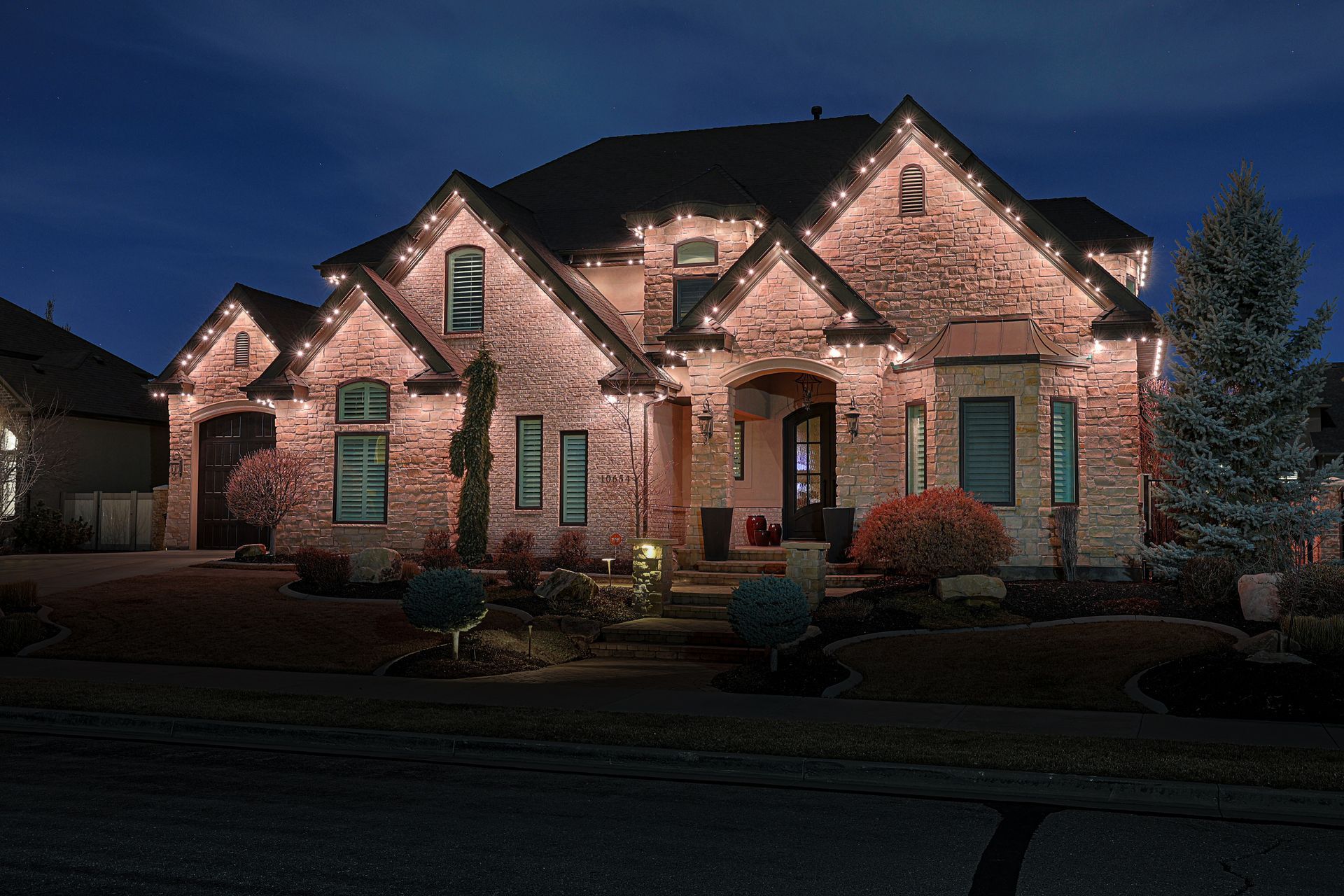 House with holiday lights at night. Brown brick, dark roof, illuminated with white lights.