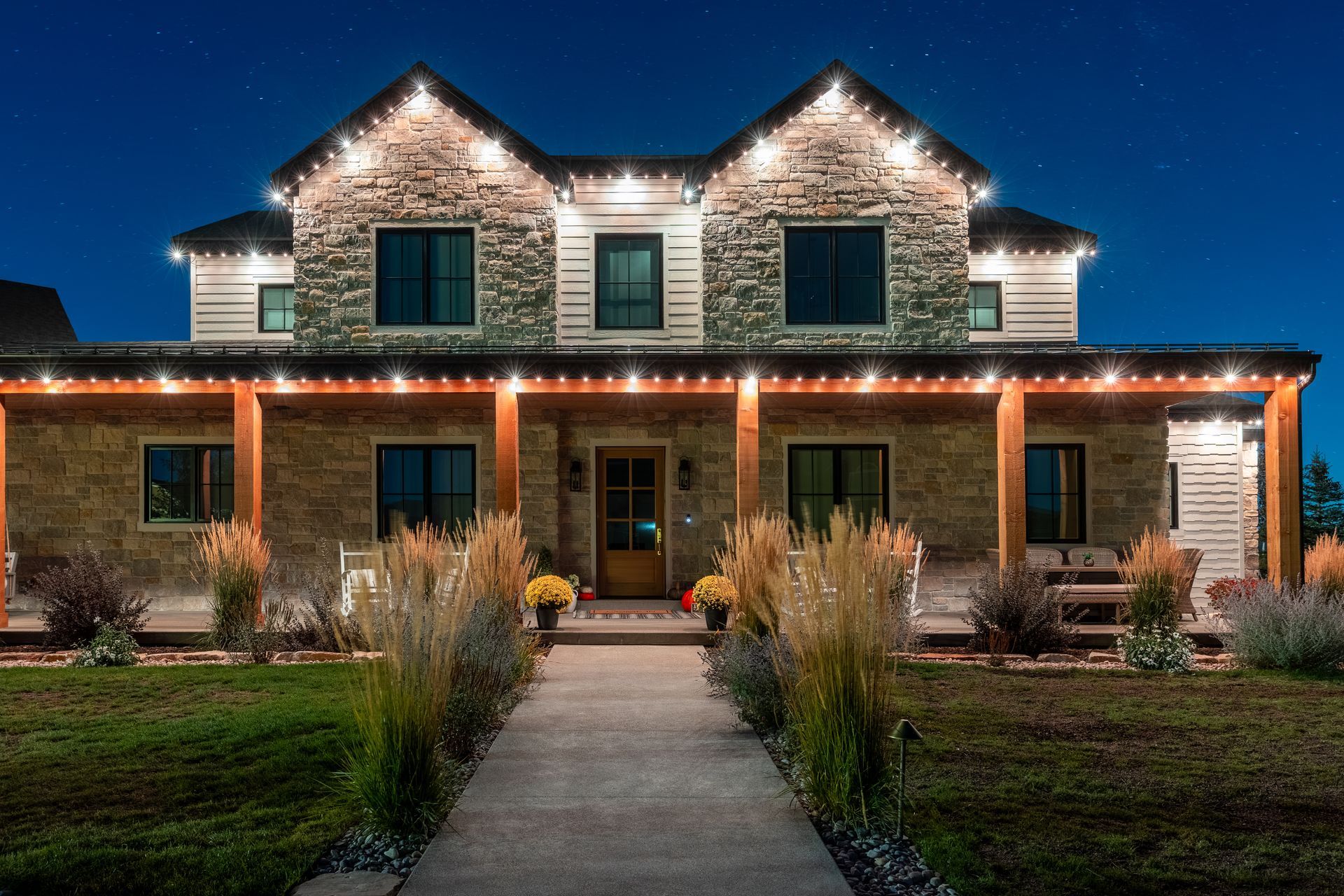 Two-story house at night, lit with white lights along rooflines and porch. Stone and light siding, walkway.