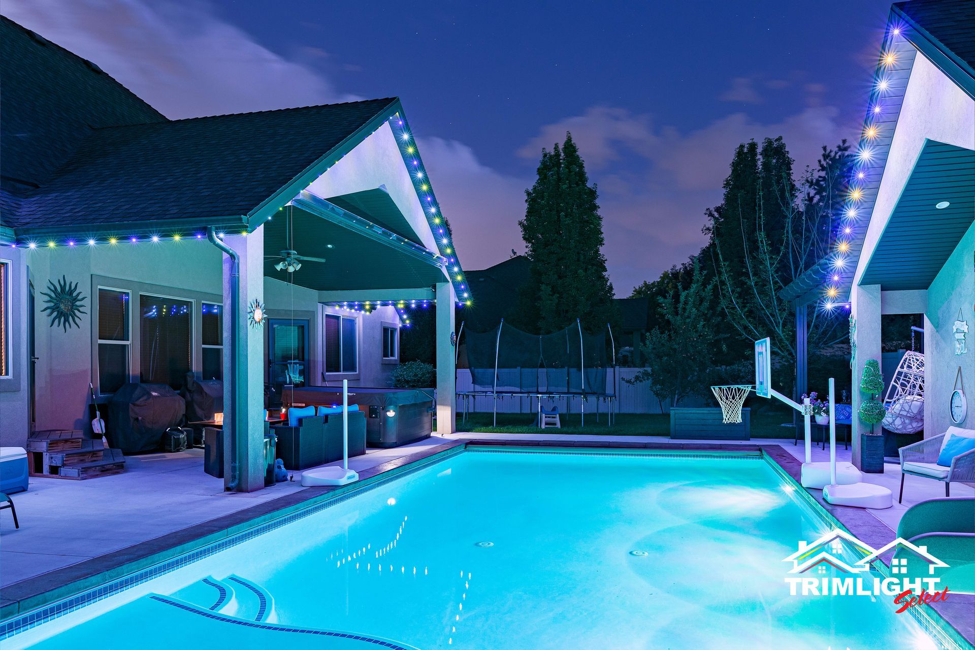 A backyard pool at night, lit by blue and white lights with a house and string lights.