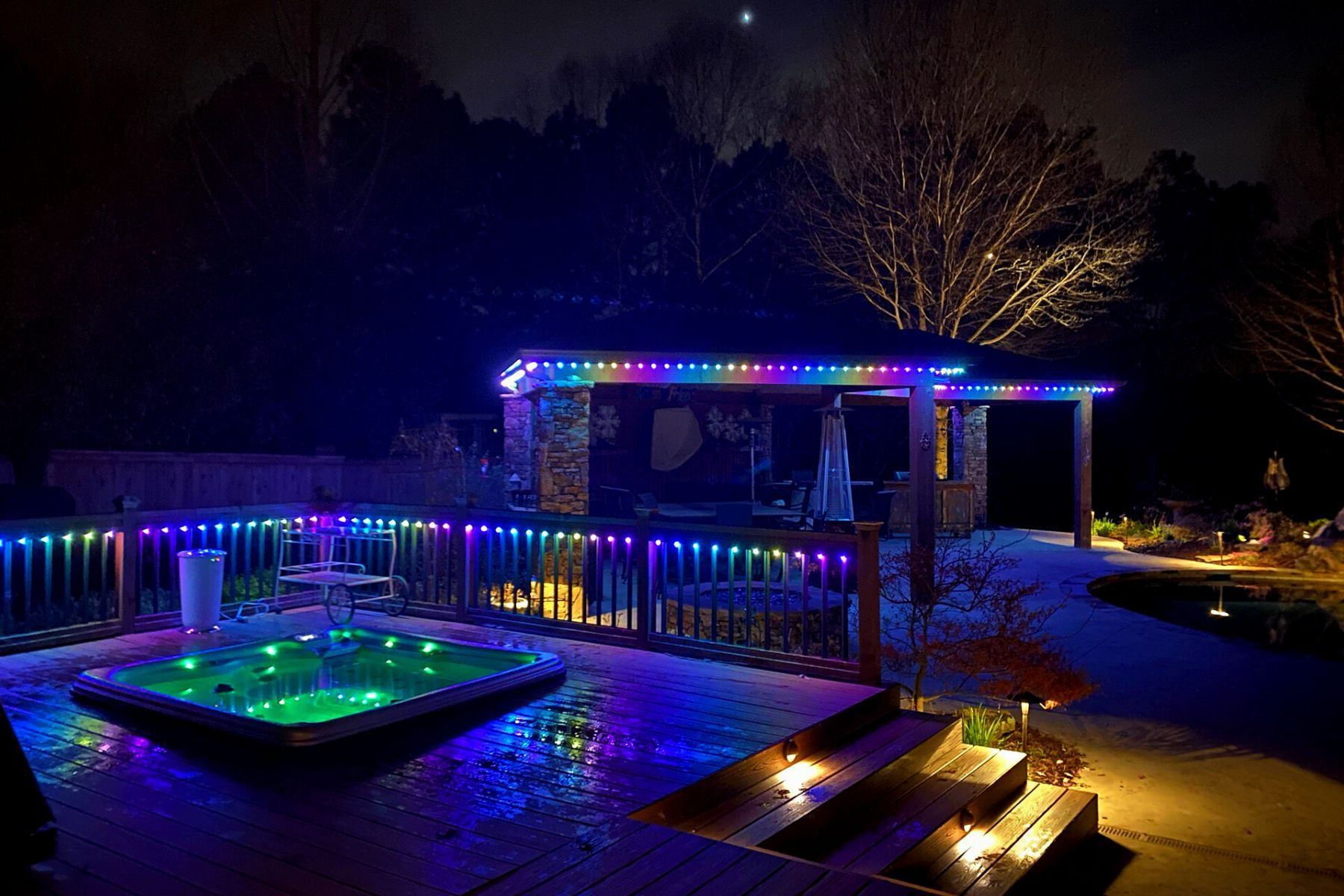 Night view of a backyard with a hot tub and gazebo illuminated by colorful LED lights.
