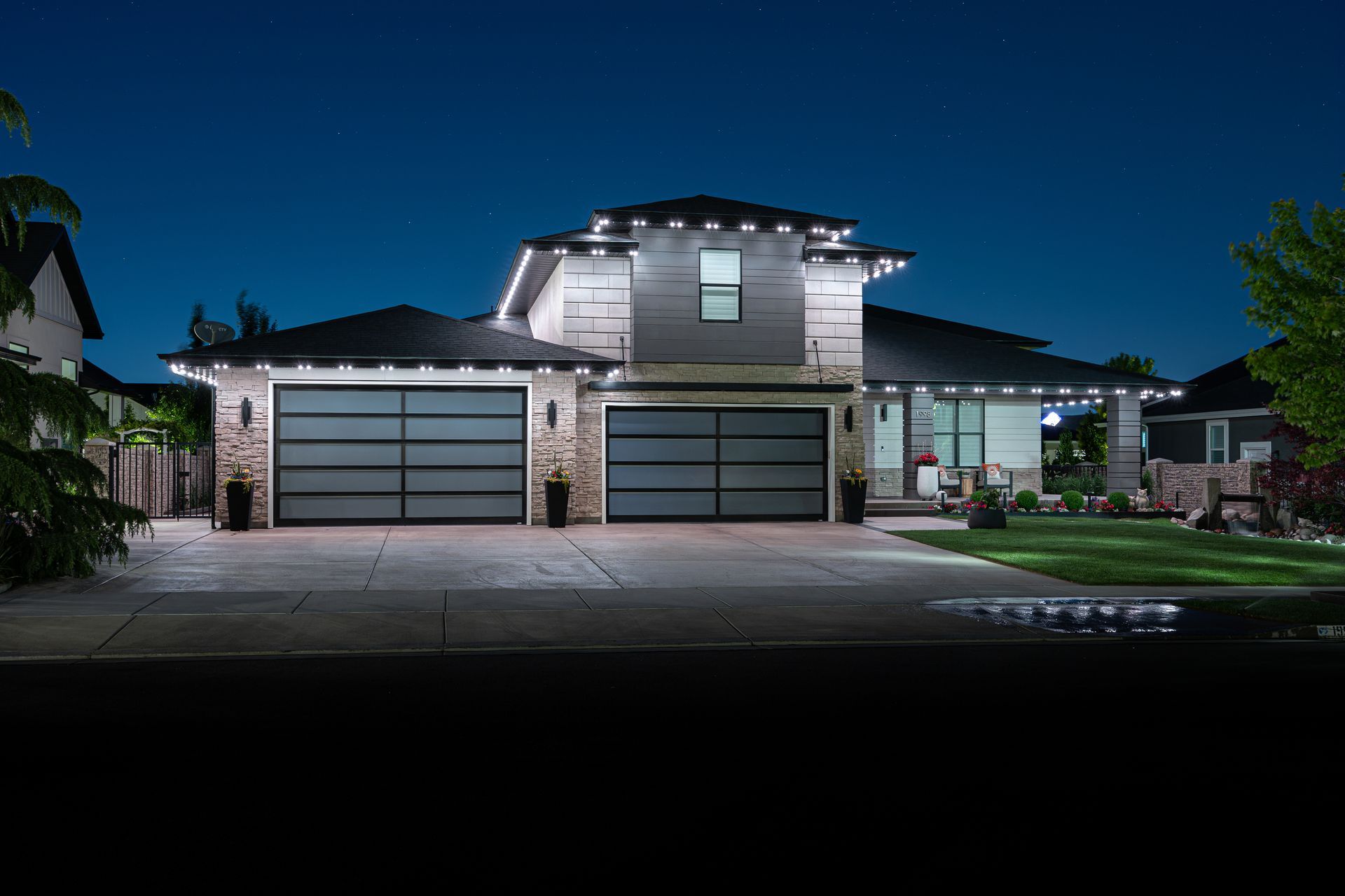 Two-story house at night with white lights outlining rooflines. Glass garage doors and lit driveway.