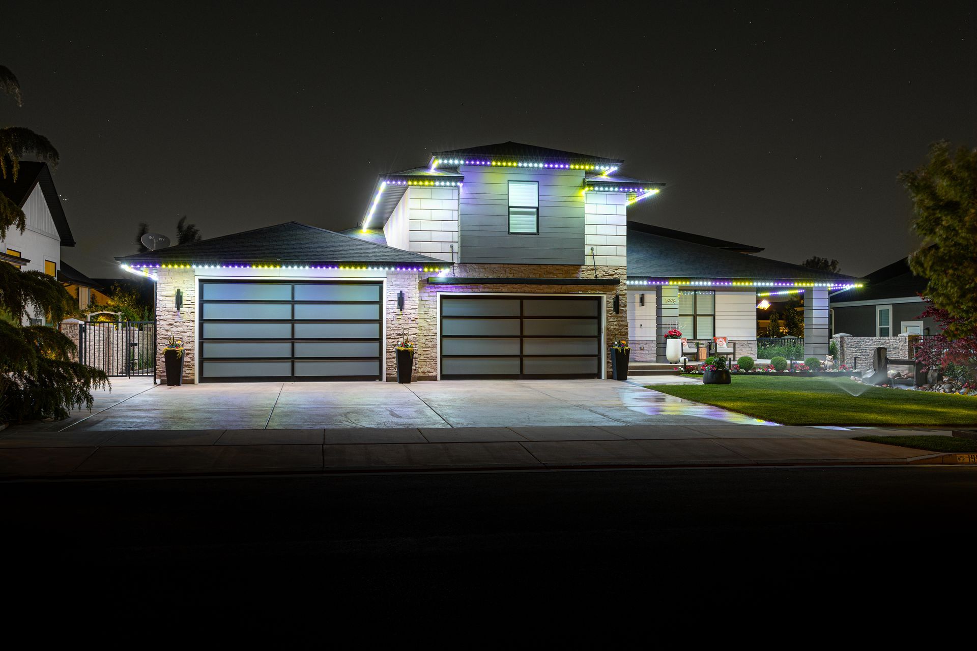 House exterior lit with colorful lights at night. Modern design with garage doors, stone accents, and manicured lawn.