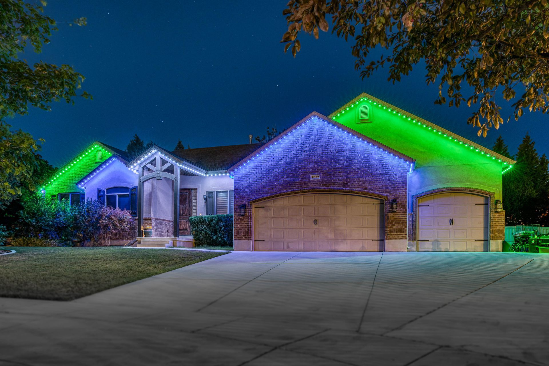 House at night, illuminated with blue and green Christmas lights. Garage, driveway.