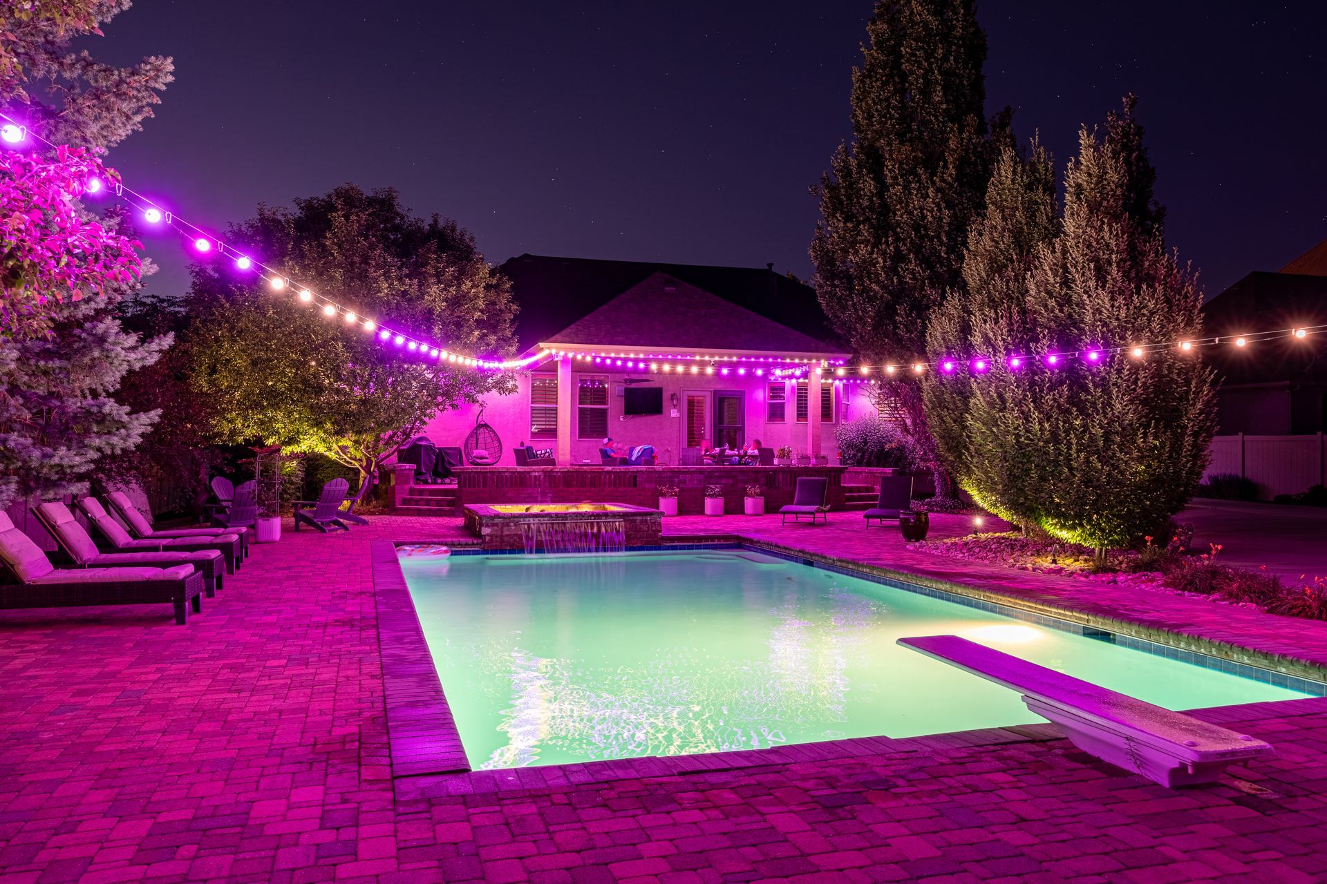 Backyard pool at night with pink and white string lights, house in background.
