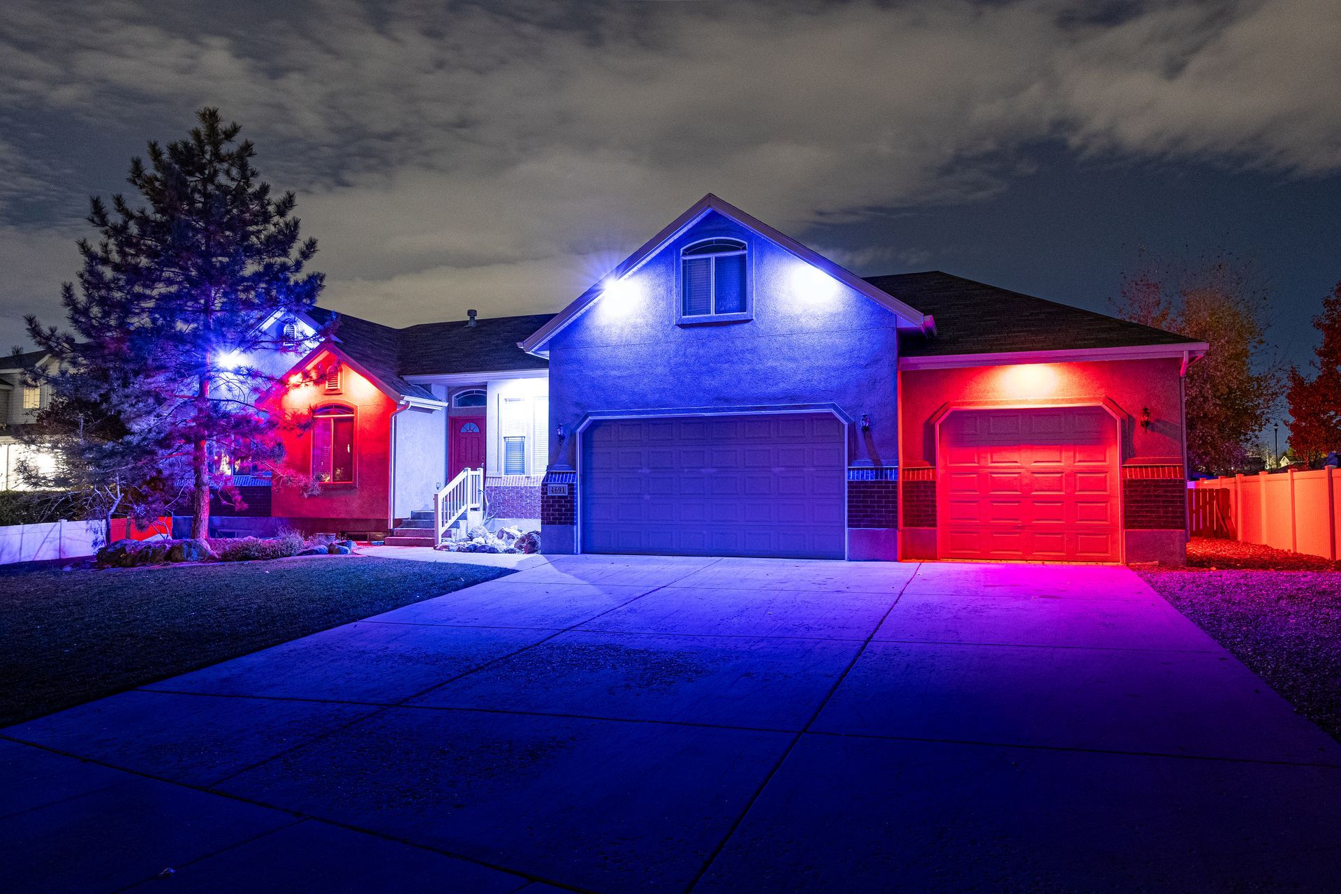 House at night lit with red and blue lights; driveway, garage, and tree visible.