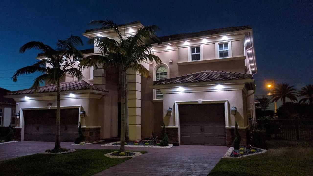 Two-story house with lights on at night, brown garage doors, palm trees, and a dark blue sky.