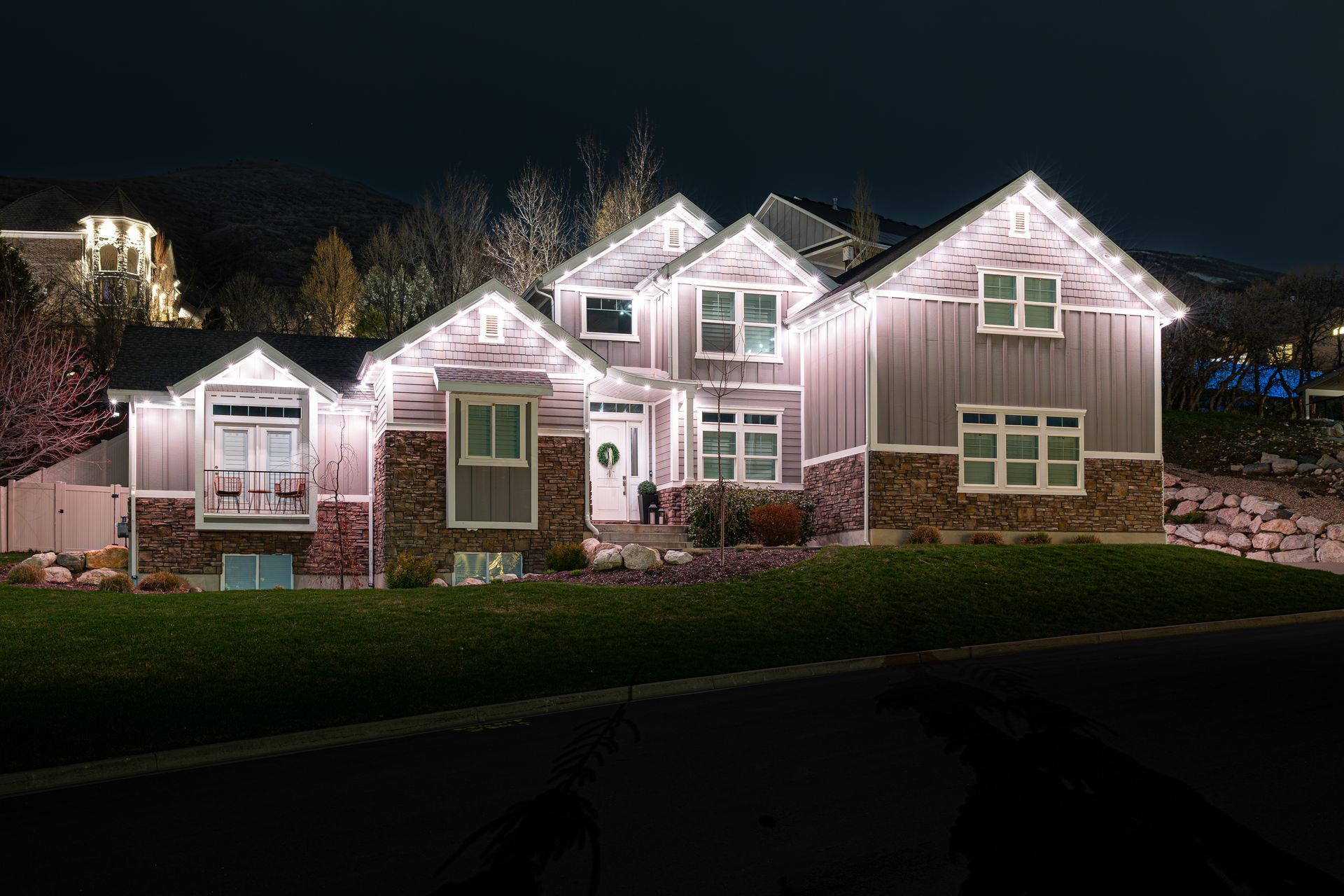 House at night with white lights outlining the roof and windows; grassy yard, dark road.