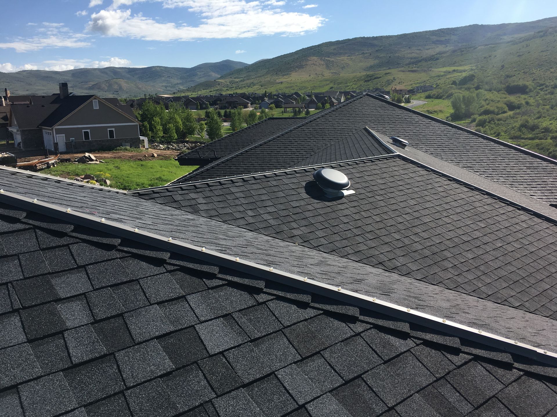 Black shingled roof with a vent and metal flashing, overlooking a landscape of homes and mountains.