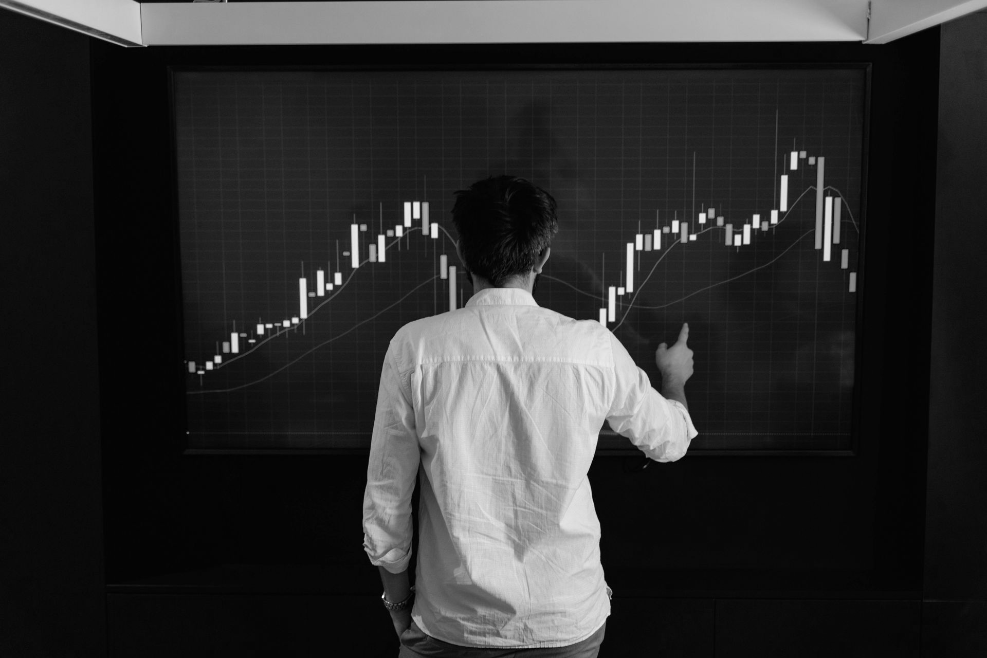 Man in white shirt analyzes stock chart on large screen, pointing.
