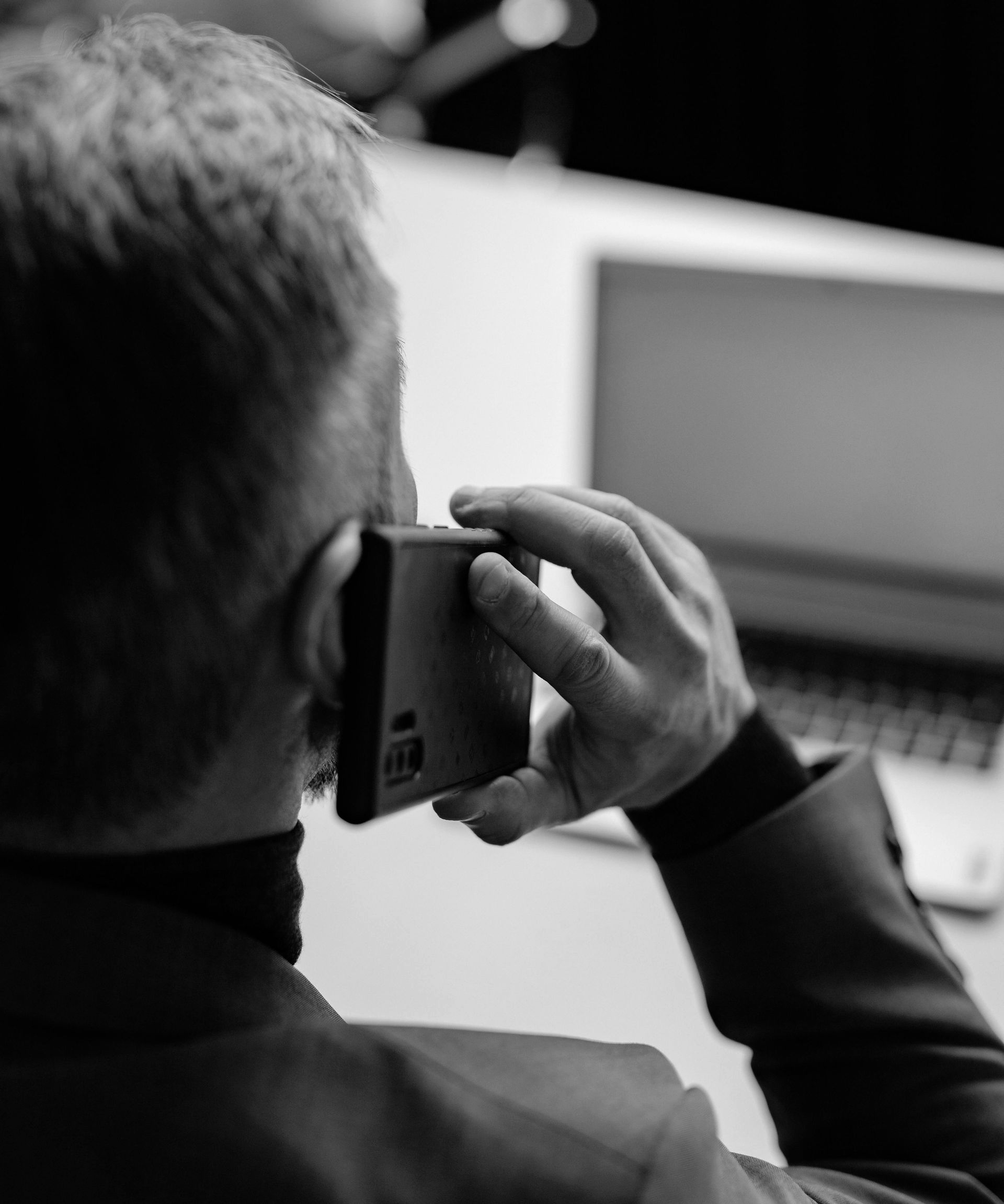 Man holding a phone to his ear, sitting at a desk with a laptop. Black and white.