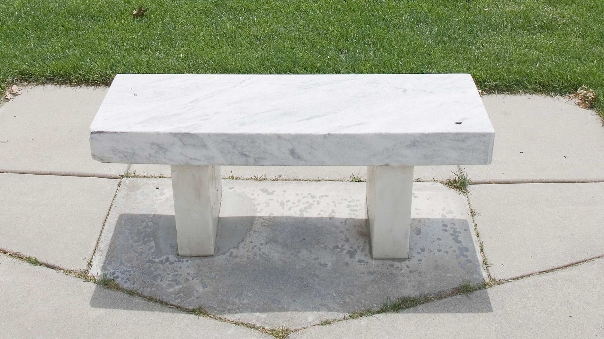 White marble bench on gray paving stones, with green grass in the background.