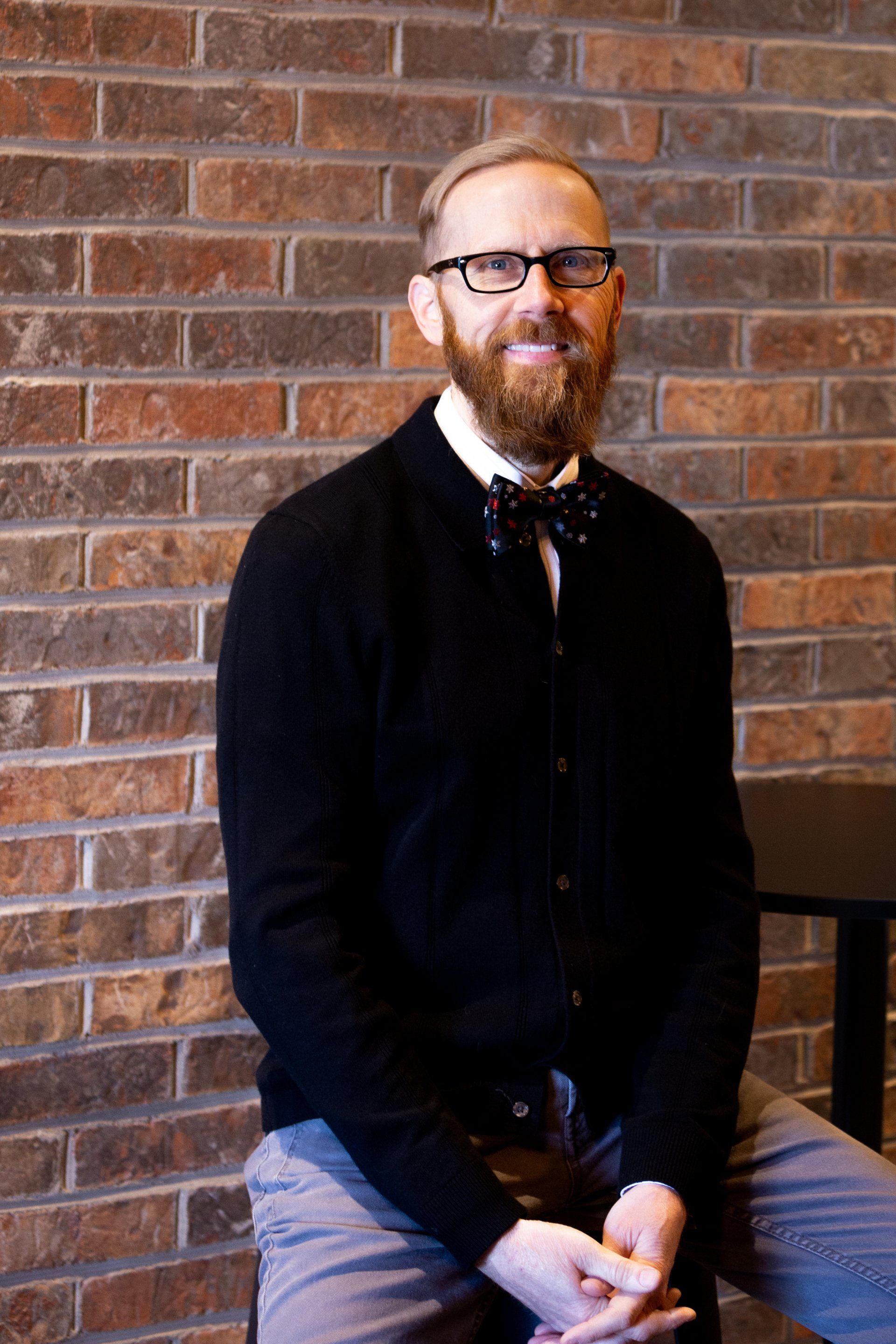A man with a beard and glasses is sitting in front of a brick wall