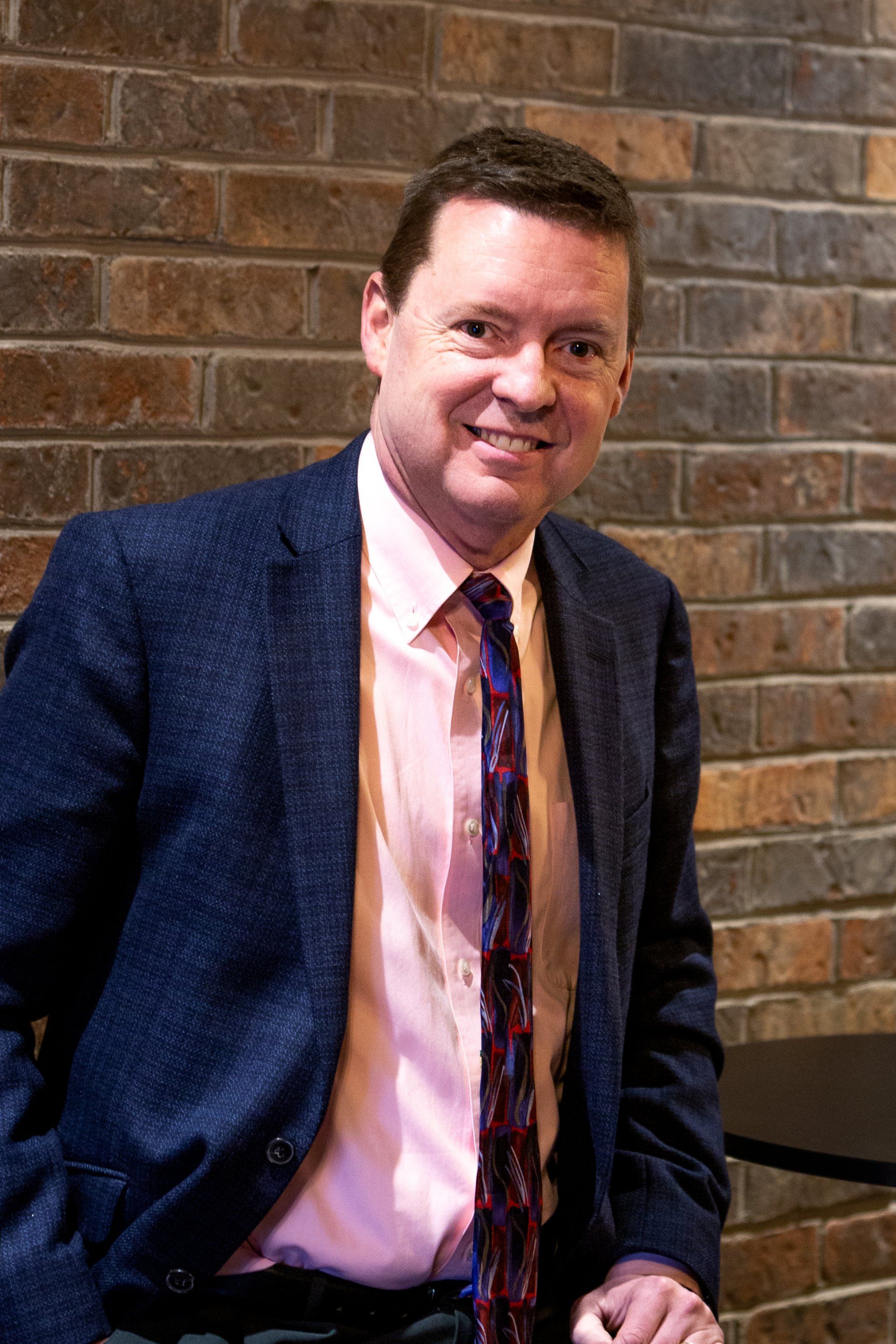 A man in a suit and tie stands in front of a brick wall