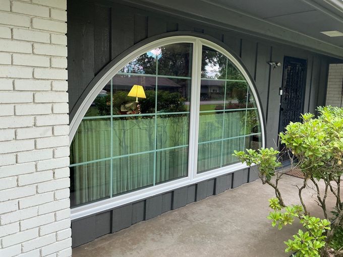 An arched, multi-pane window sits on a gray-sided home exterior next to white brick, with a green plant in the foreground.