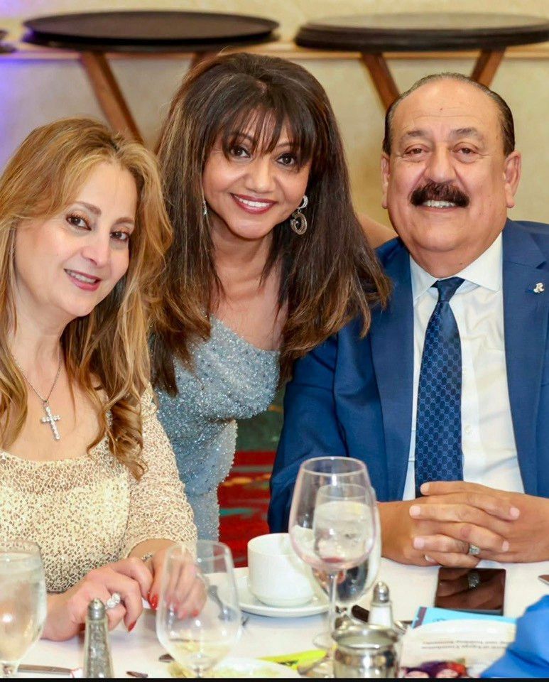 Three people smiling at a table; woman in blue gown, man in blue suit, woman in gold top.