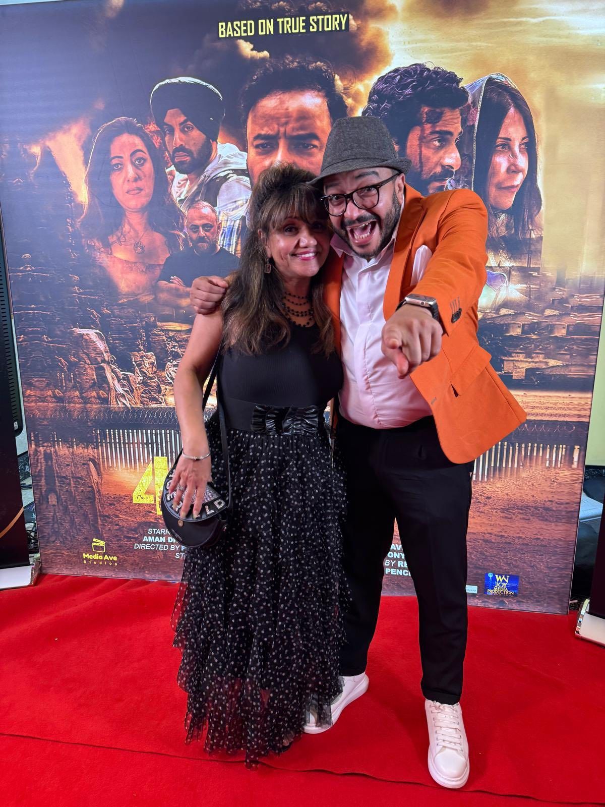 Two people posing on a red carpet in front of a movie poster. The man points enthusiastically.