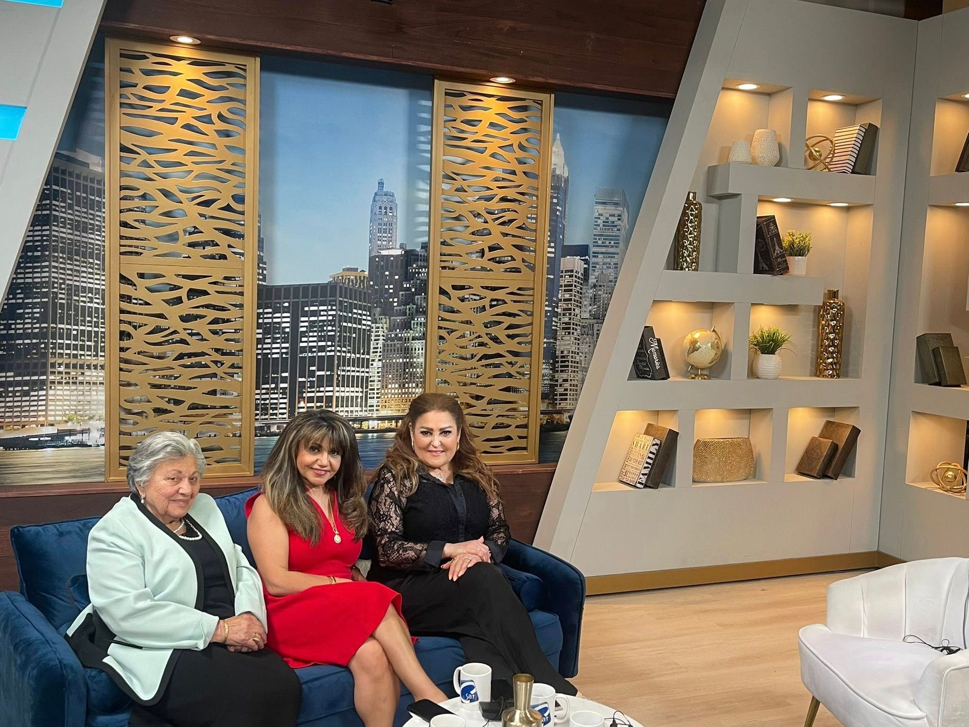Three women seated on a blue sofa in a studio. The background features a city view and decorative wall panels.