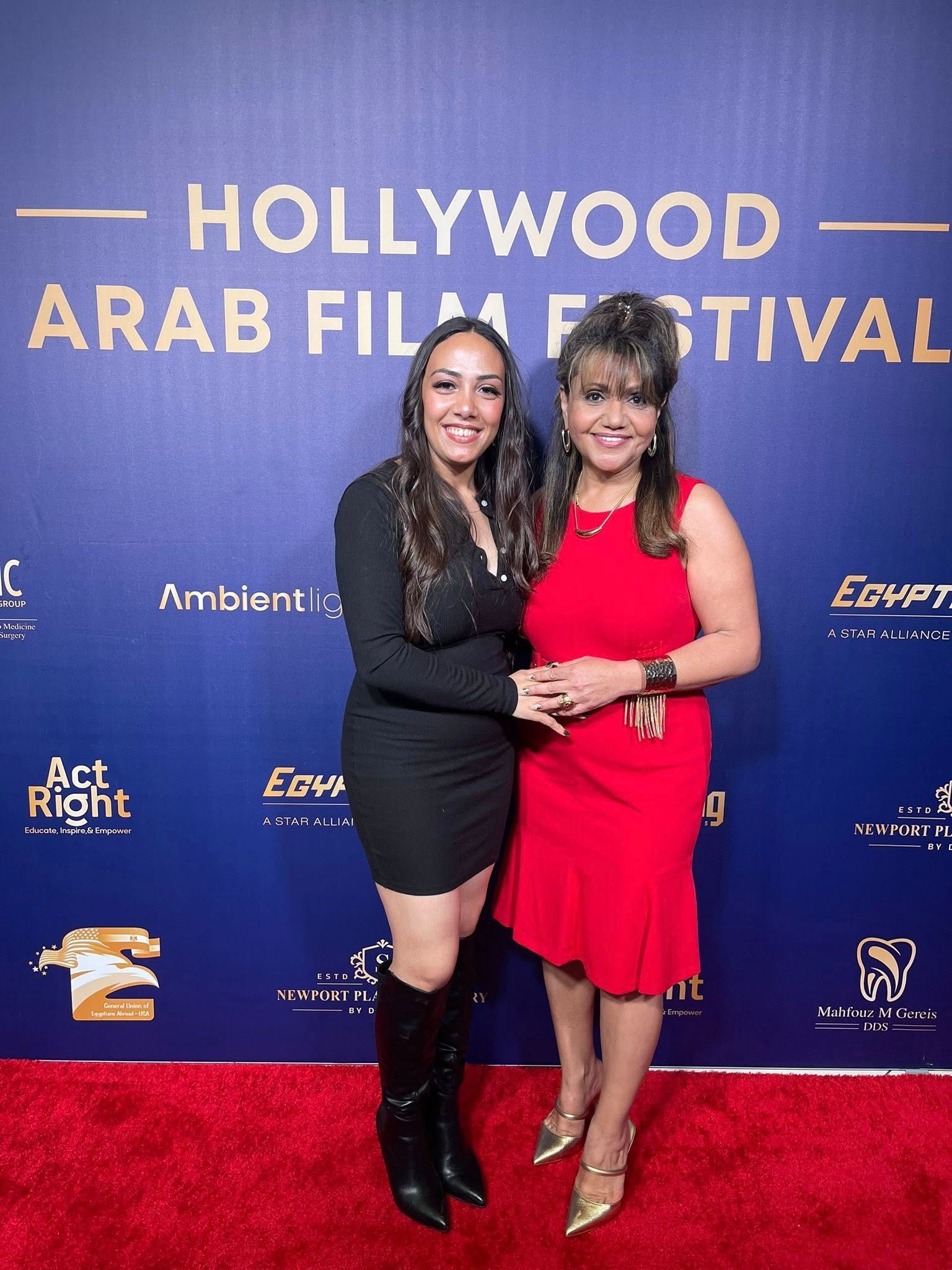Two women pose on a red carpet at the Hollywood Arab Film Festival; one wears a black dress, the other a red dress.