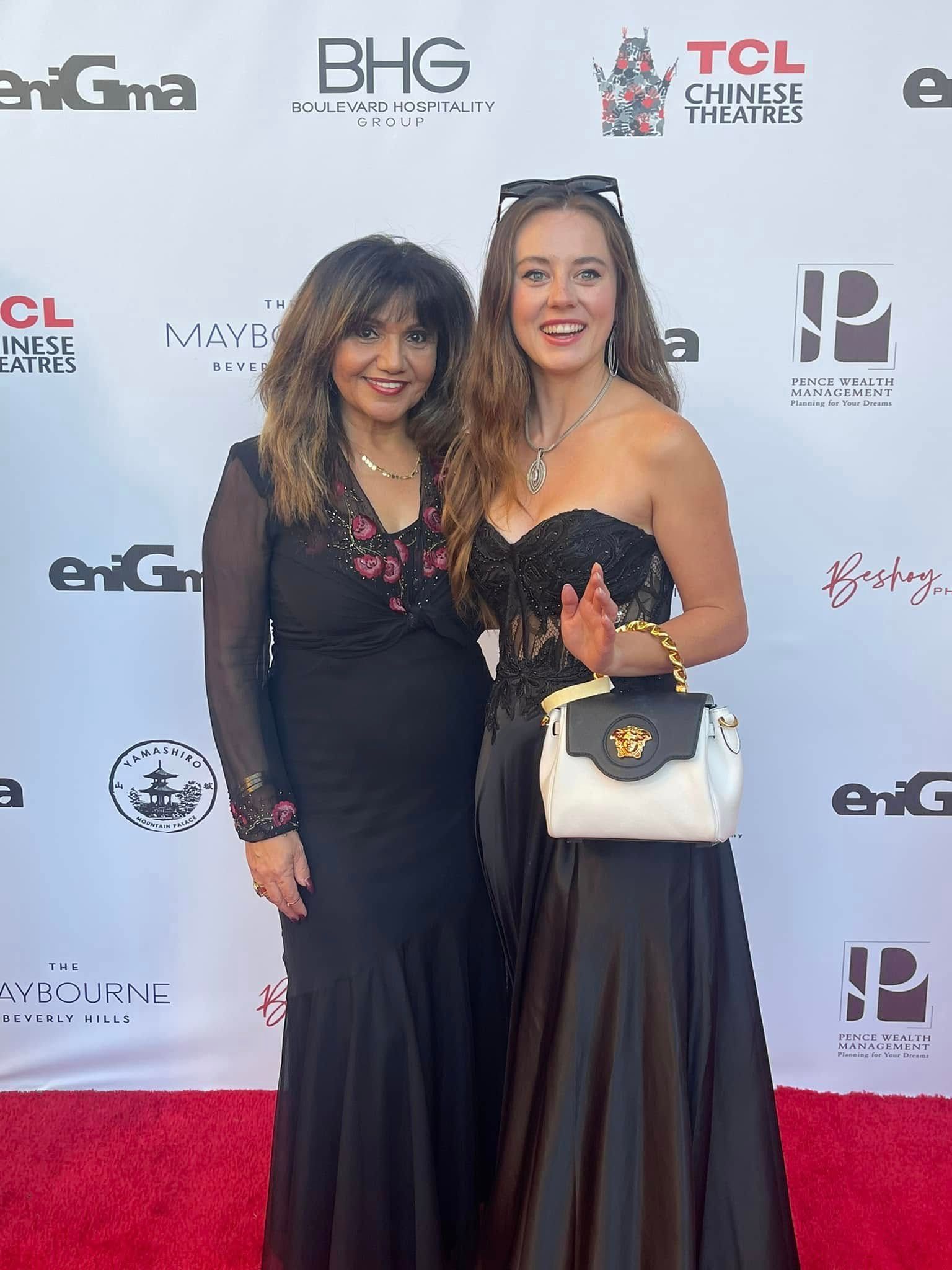Two women pose on a red carpet. The woman on the left wears a black dress with pink accents. The other woman wears a strapless black gown and holds a white purse.
