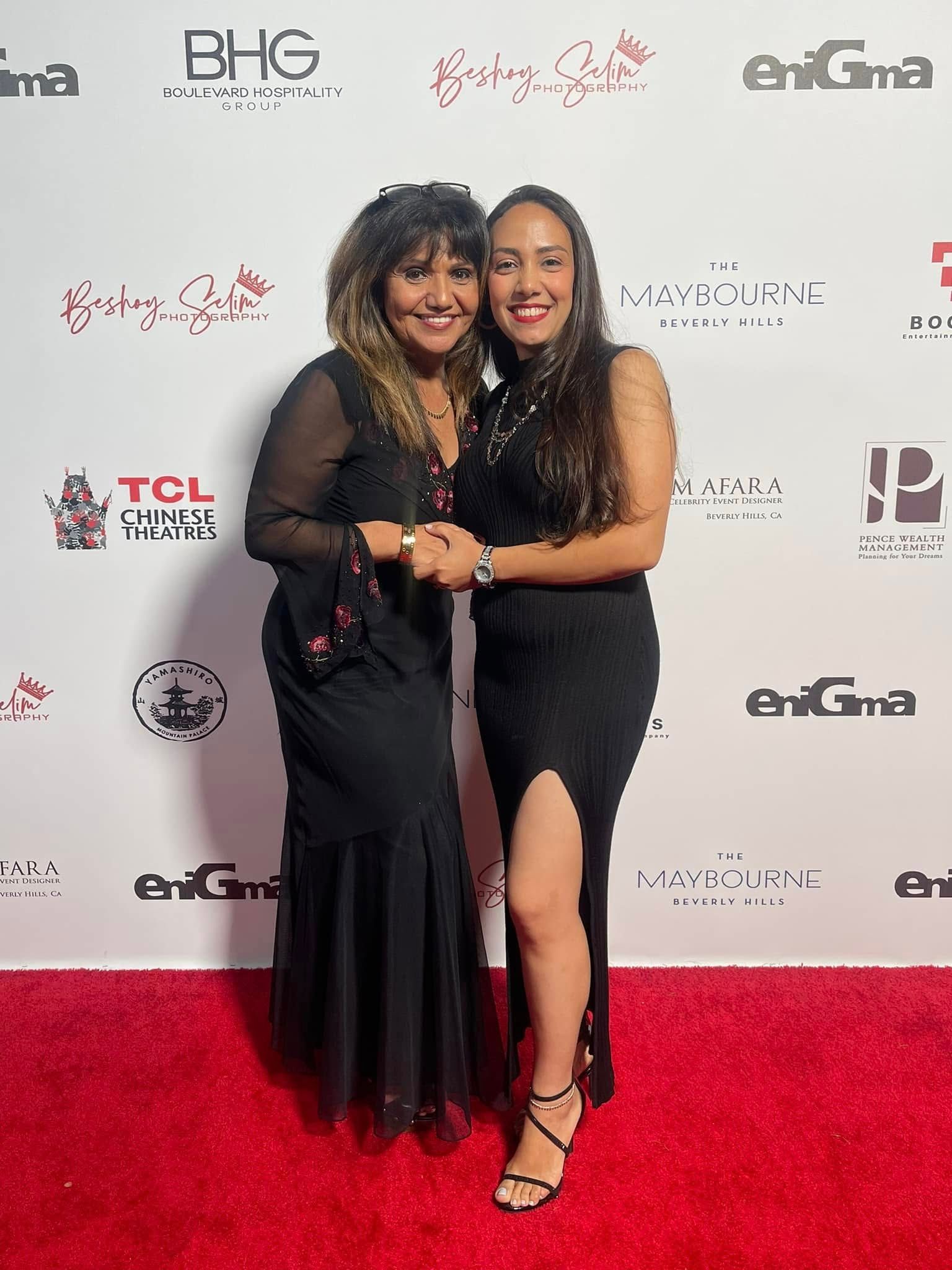 Two women in formal black dresses pose on a red carpet with event logos in the background.
