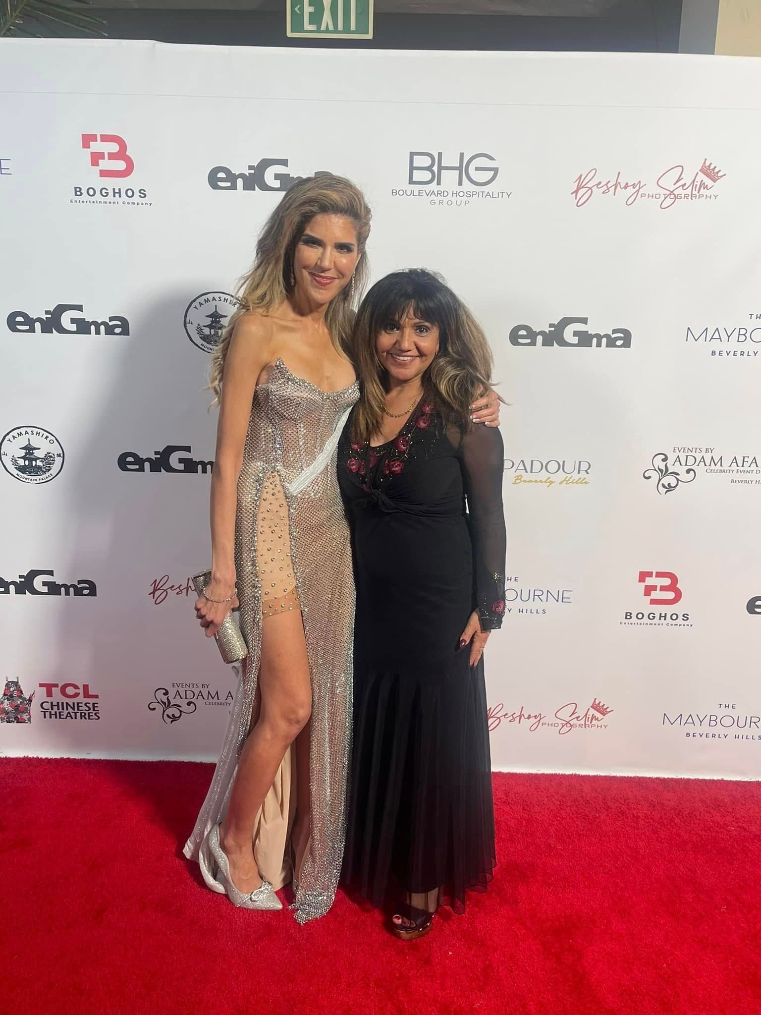 Two women pose on a red carpet. One in a sequined gown, the other in a black dress, both smiling.