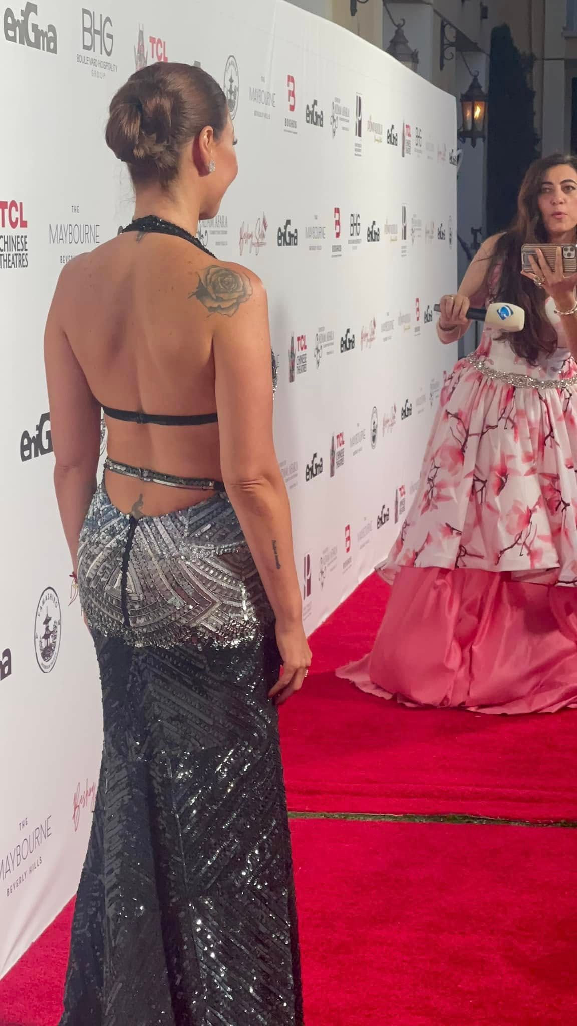 Woman in a sequined gown poses for a photo on a red carpet. Back is exposed. A person takes photos nearby.