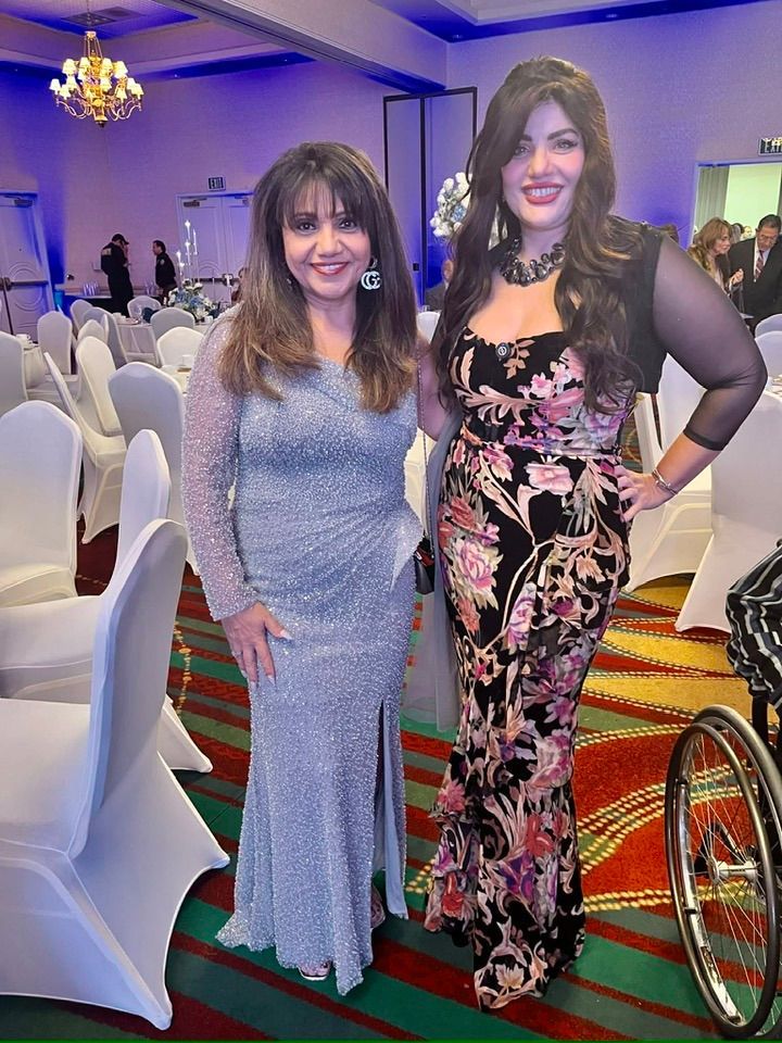 Two women pose at a formal event, one in a sparkling silver gown, the other in a floral dress and black bolero.