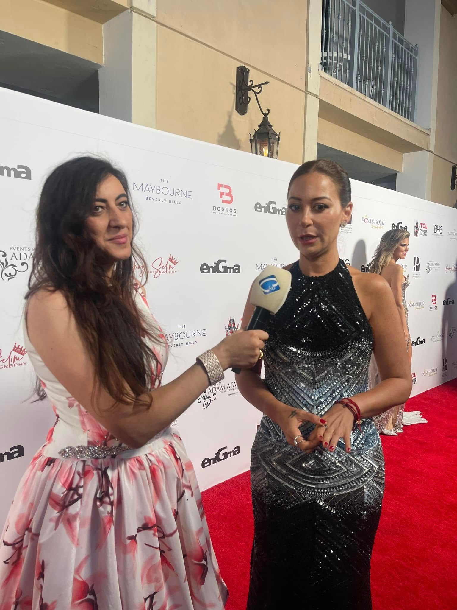Woman being interviewed on a red carpet; interviewer holds a microphone. Buildings are in the background.