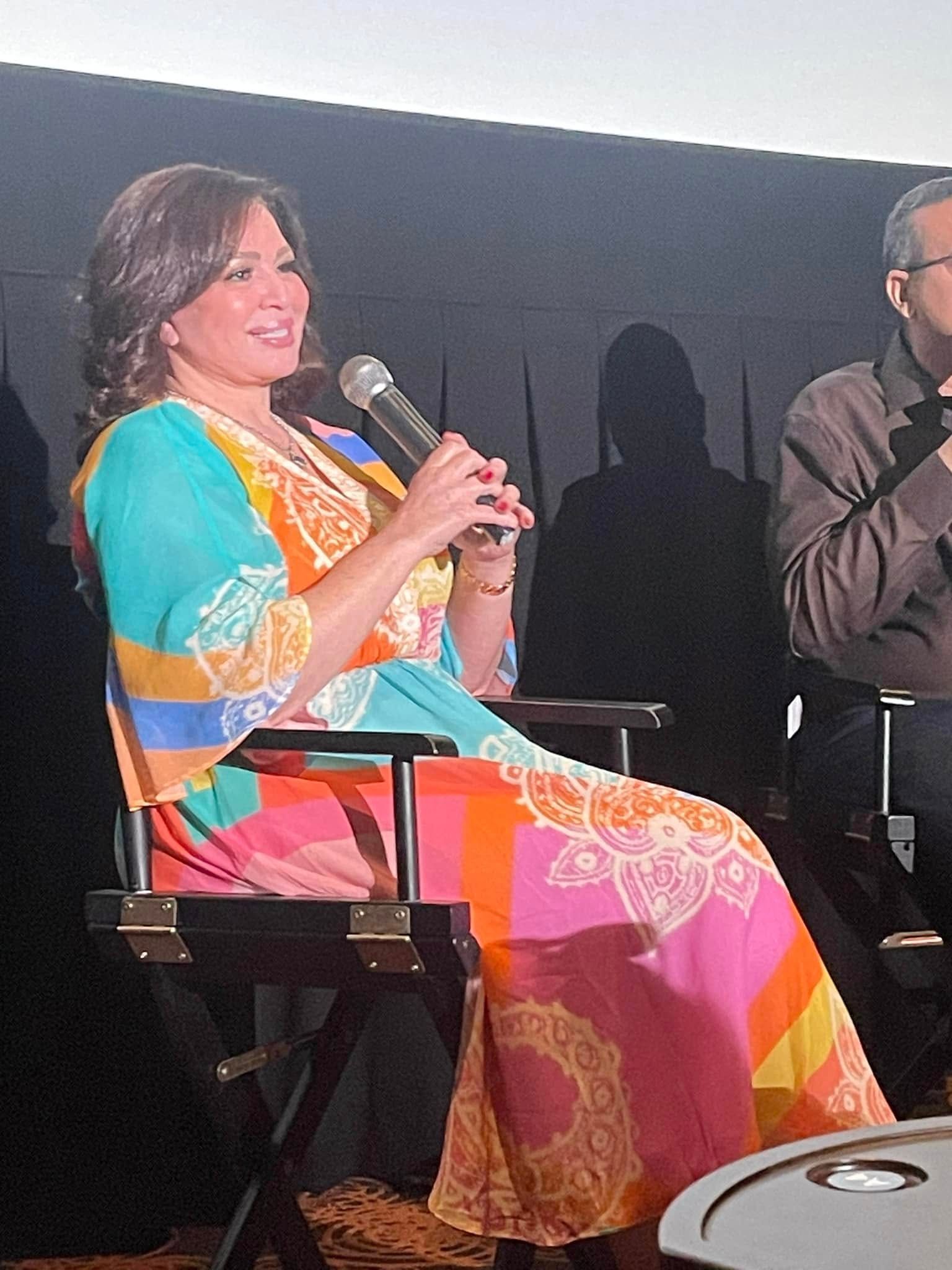 Woman in colorful dress speaking into a microphone, seated on a director's chair at an event.