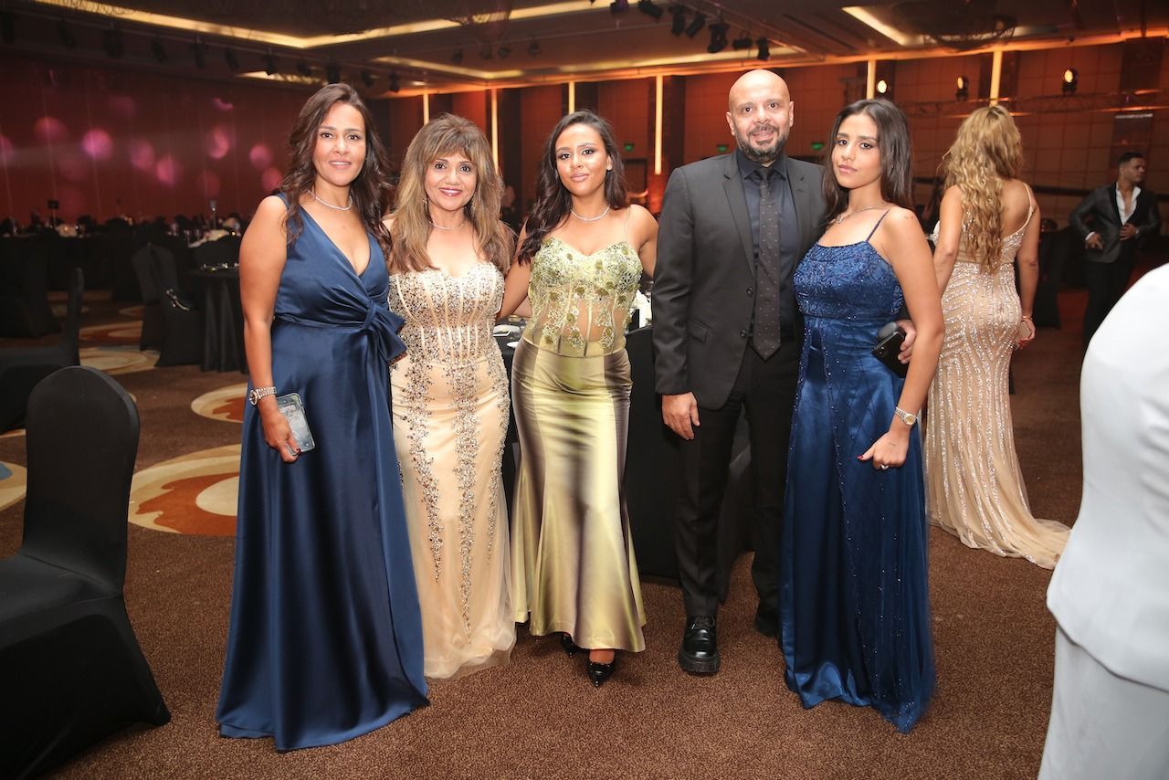 Group of people in formal attire pose at a gala. Dresses range in colors, room has dark decor.