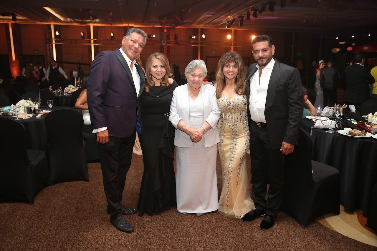 Five people pose at a formal event, including a woman in a gold gown and another in a white dress.