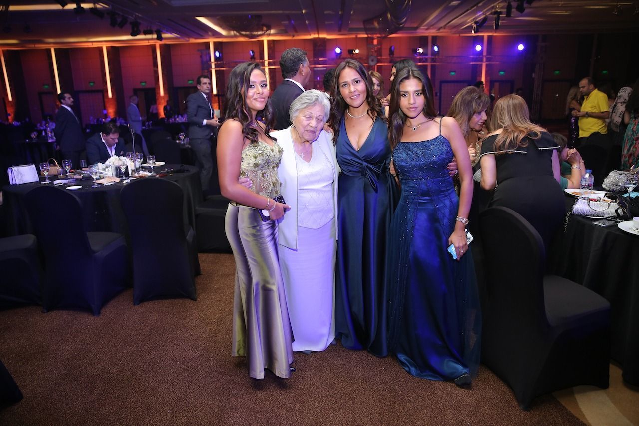 Four people pose for a photo at a gala. Three women in gowns surround an elderly woman in white.