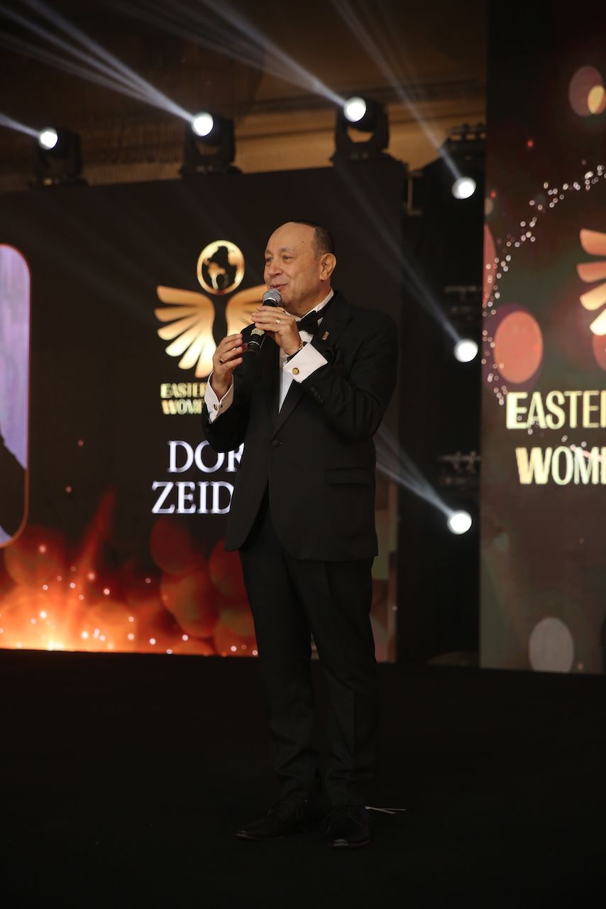 Man in a tuxedo speaking into a microphone on a stage with a logo and lights.