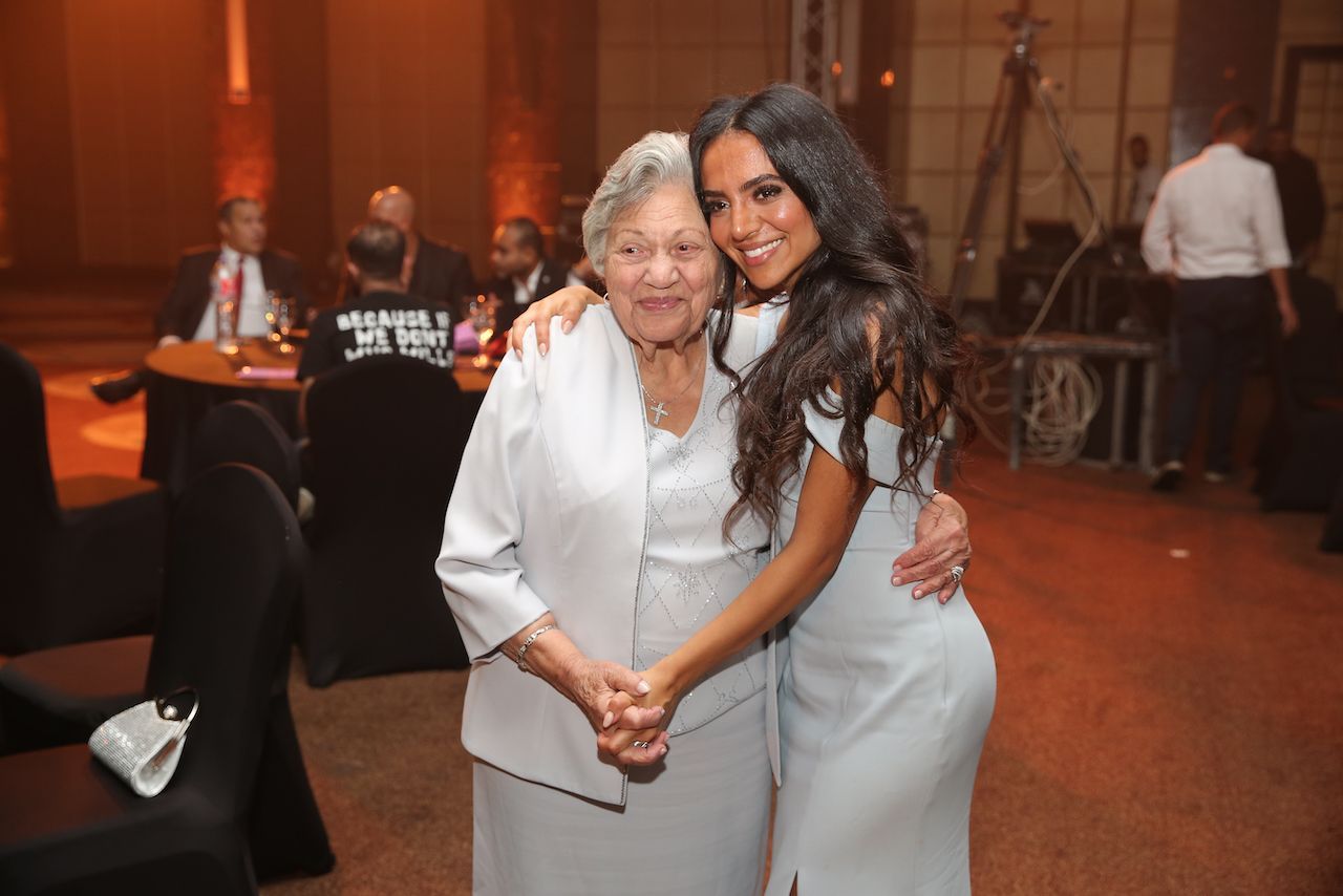 Woman embraces older woman; both smile at a formal event, lit in warm tones.