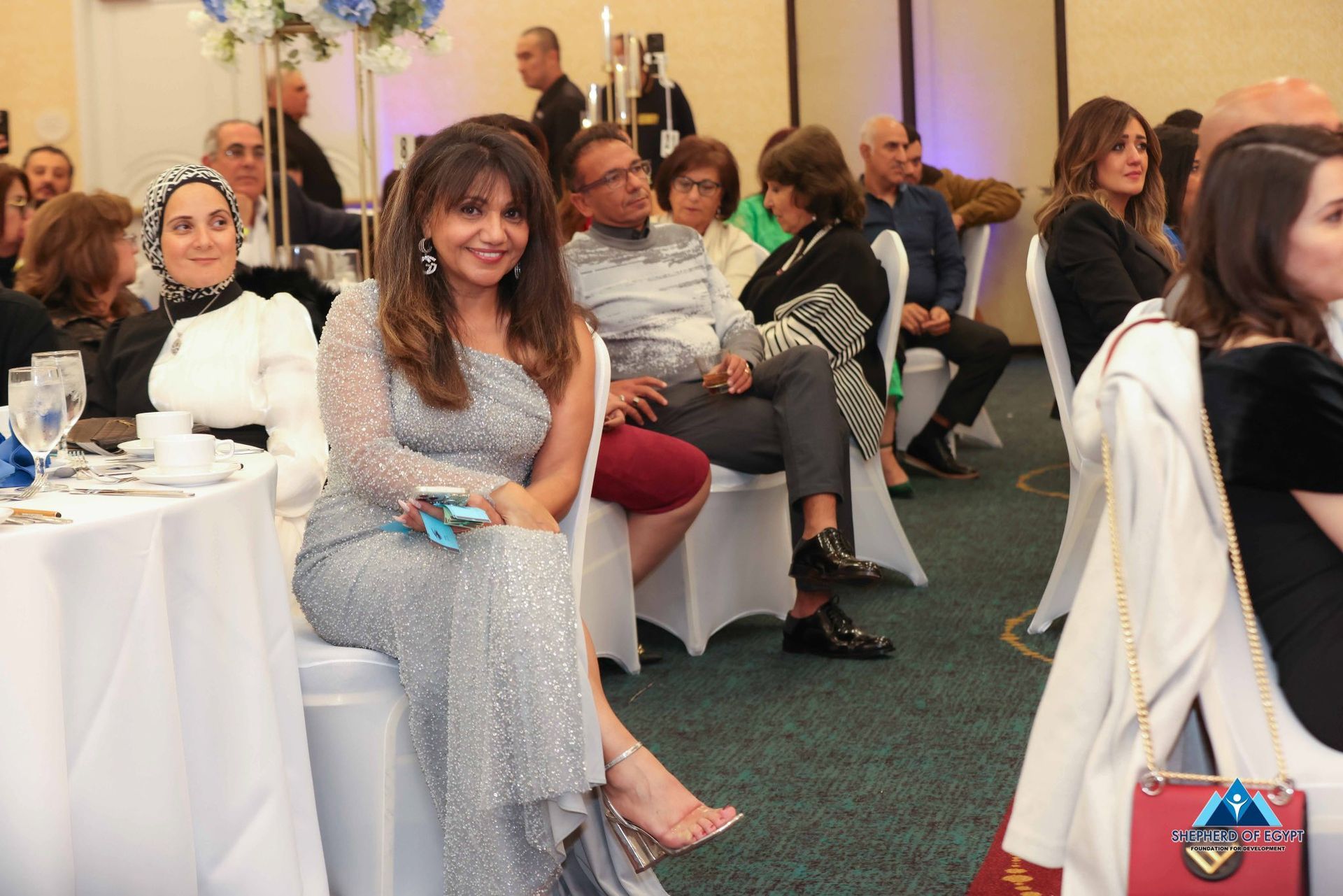 Woman in silver dress seated at a table, smiling. Guests at event in a brightly lit room.
