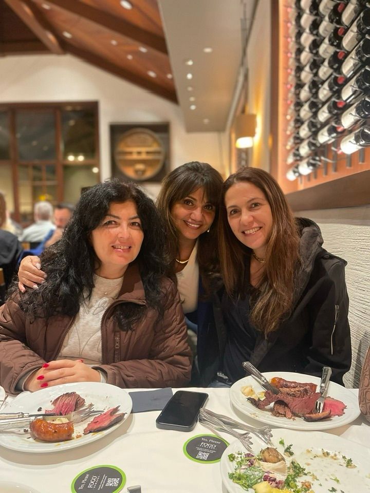 Three women smiling at a restaurant table with plates of food. A wine rack is in the background.
