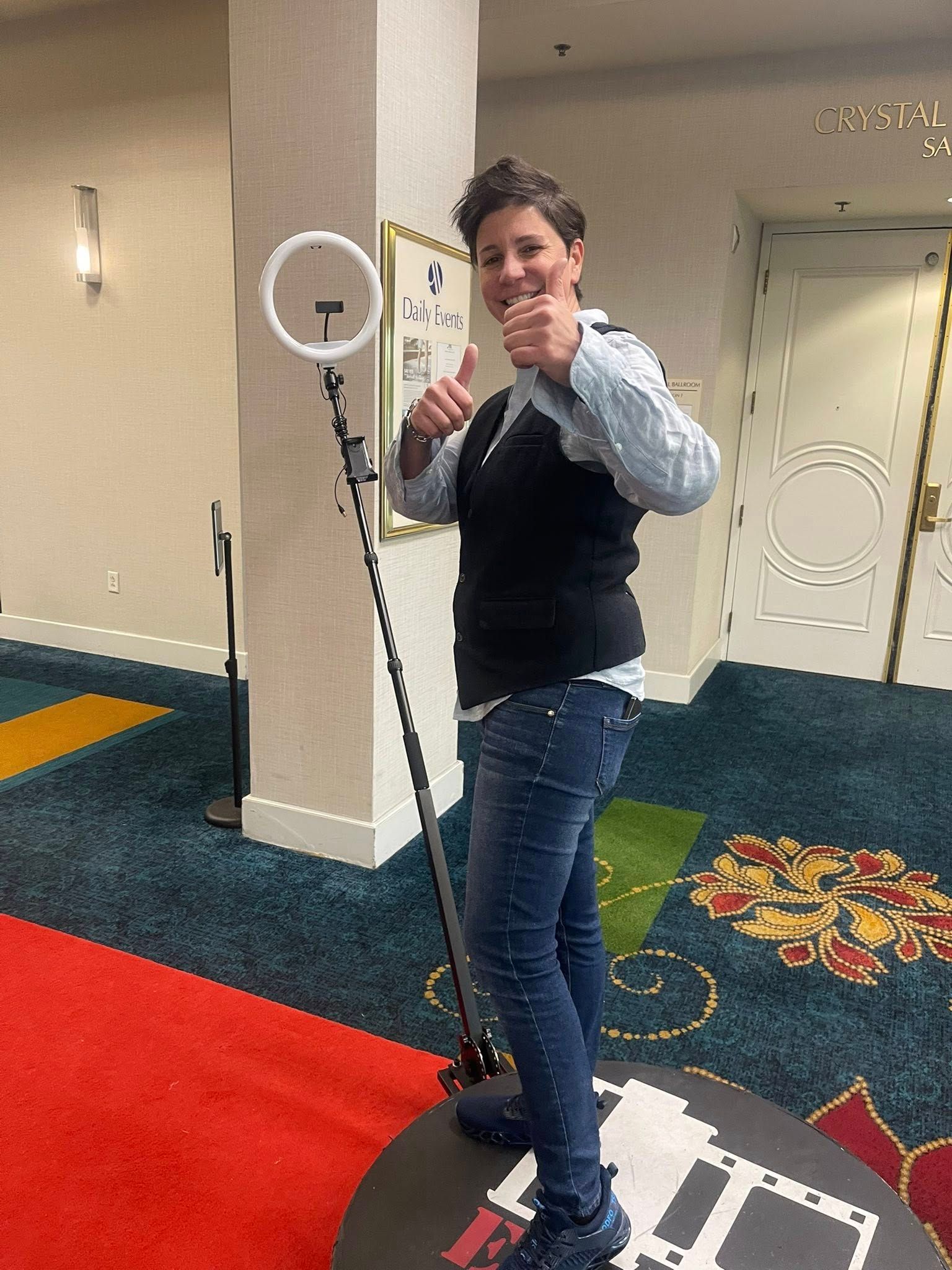 Person standing on a rotating photo booth platform, giving thumbs up. Wearing dark vest, jeans, and sneakers.