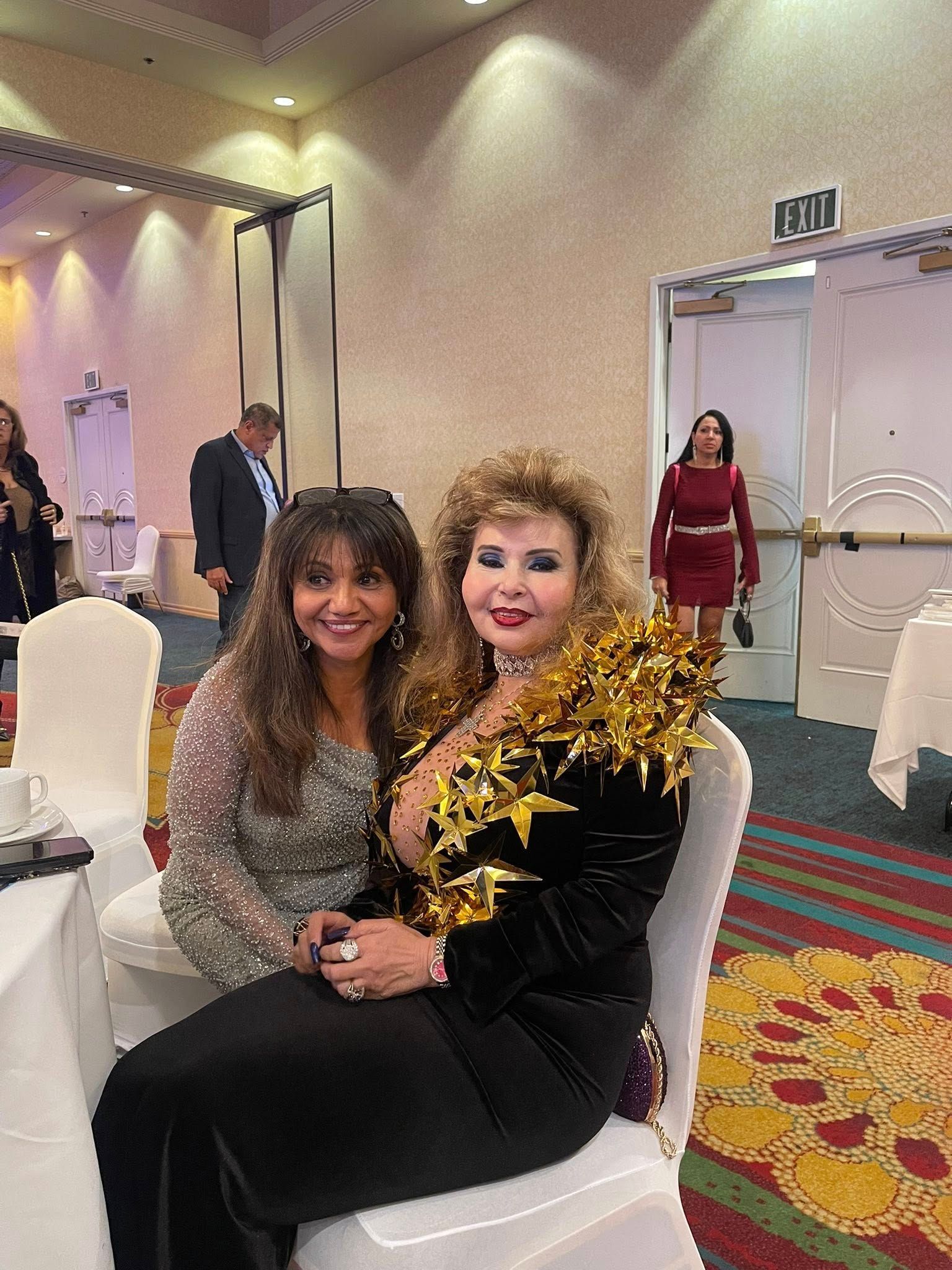 Two women seated at a table, one in a sequined top, the other in black with gold embellishments, at an event.