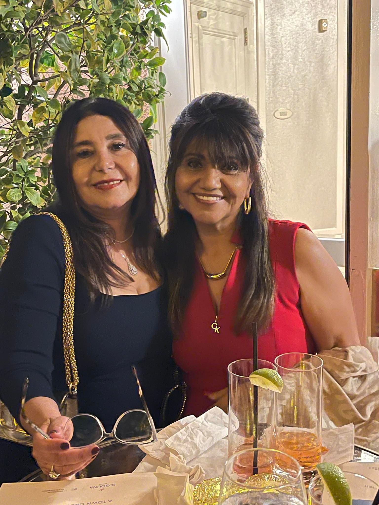 Two women smiling at a table, one in red, one in black, drinks and greenery visible.