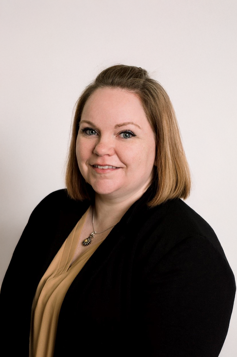 A person with shoulder-length brown hair, wearing a black cardigan over a tan top and a pendant necklace, smiling.